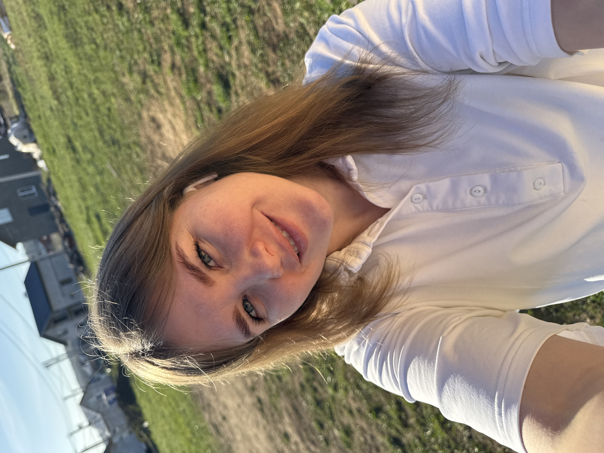 A light-skinned young woman with straight, light brown hair poses for a selfie outside on a sunny day in Ферма, Russia. She wears a white collared shirt with buttons. Buildings are visible in the background to the left, beyond a grassy area. The ground is covered in green grass.