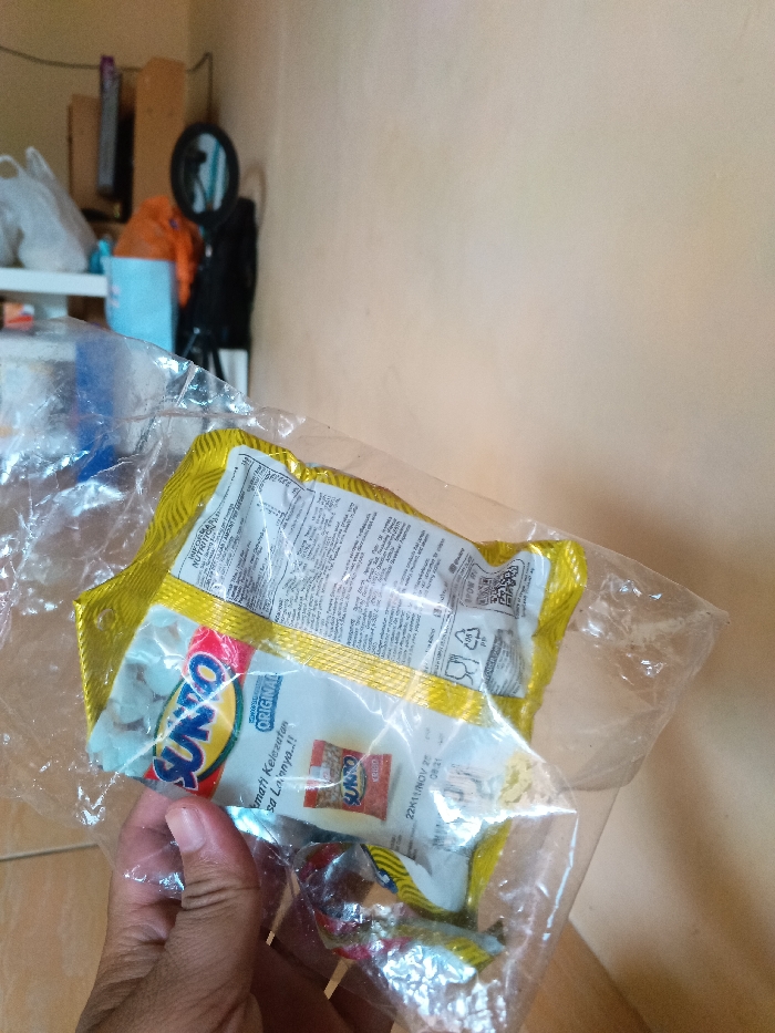 The image captures a close-up of a hand holding a clear plastic bag filled with snack packages against an indoor background, likely in Cijeungjing, Indonesia.

In the foreground, a person's hand, with a medium skin tone, grips a transparent plastic bag. Inside the bag, several small, individually wrapped snack packages are visible. The most prominent package is white and yellow, featuring the brand name "SUNTO" in red and blue letters, along with the word "ORIGINAL". Below this, the phrase "Nikmati Kelezatan Rasa Lainnya..!!" (Enjoy the taste of others..!!) is printed. A small image of a snack is also visible on this package. Text that appears to be a production or expiry date "22K11 NOV 25 08:21" is also present. Behind this package, another yellow package shows partially visible text, including "INFORMASI NUTRITION" and what appears to be recycling symbols. A small, red-wrapped candy or lollipop with a stick is also present at the bottom of the bag.

The background reveals a light-colored, possibly beige or light orange, wall on the right side. To the left, various household items are stacked or placed on shelves, including a white plastic bag, an orange object, a light blue container, and a dark circular object that resembles a mirror or a microphone pop filter on a stand. The scene suggests a casual indoor setting, possibly a storage area or a room within a home. No specific time of day or weather conditions are discernible.