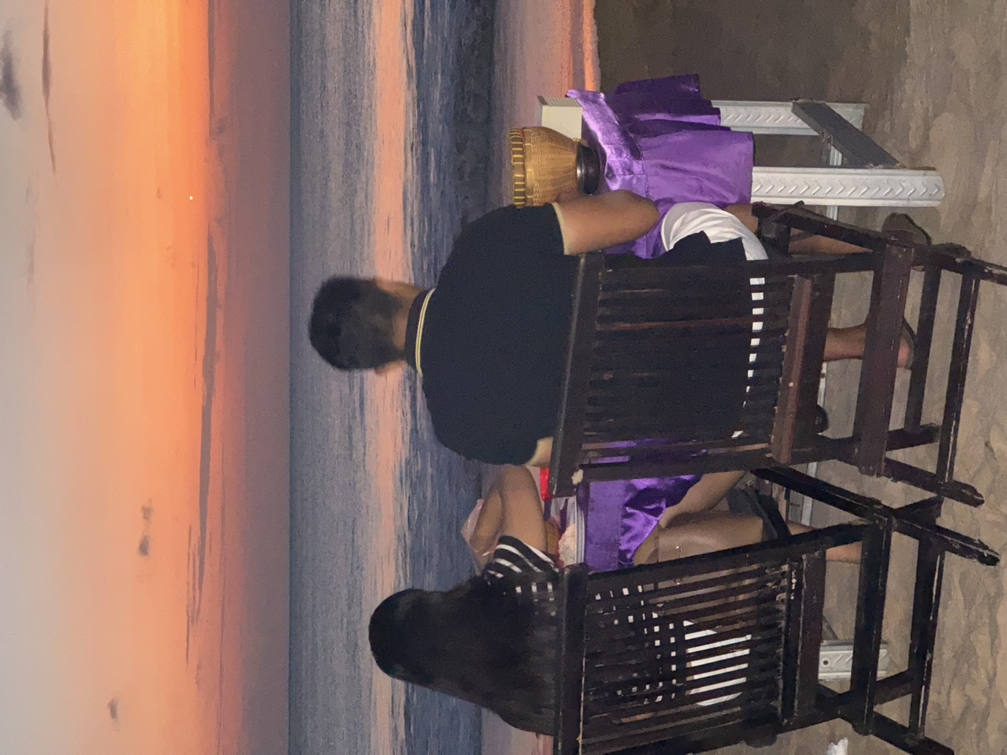 This image depicts a couple dining on a beach in Kuta Selatan, Indonesia, at dusk. The sky is ablaze with the warm hues of a sunset, transitioning from orange to pink, with a few wispy clouds scattered across the horizon. The ocean stretches out before them, its surface reflecting the fading light.
The couple is seated at a table covered with a vibrant purple tablecloth. The woman, with long dark hair, faces the table, her hands occupied with her meal. She wears a black and white striped shirt. The man, facing away from the camera, is dressed in a black polo shirt with a yellow collar trim and white shorts. On the table, a woven basket sits beside a white box, suggesting a meal or offering.
Two dark wooden chairs, designed for outdoor use, frame the scene. The table itself is supported by white pillars with an embossed pattern, typical of beachside dining setups. The sandy beach forms the foreground, and the gentle waves of the ocean create a serene backdrop. The overall impression is one of a tranquil evening meal by the sea.