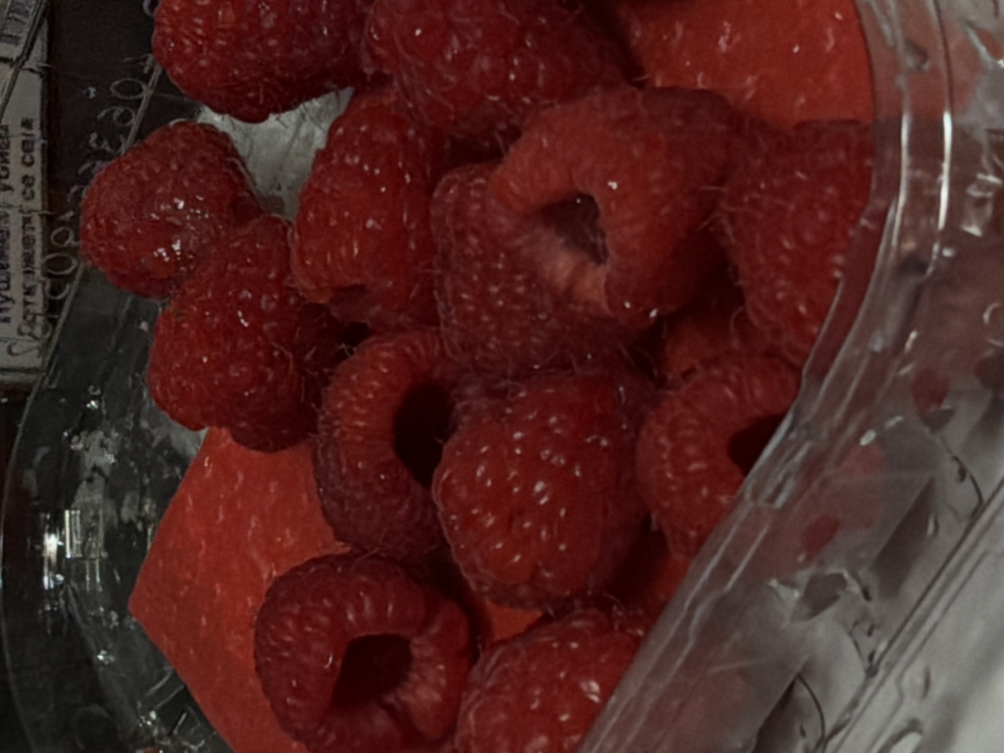 A close-up, overhead shot shows a clear plastic container filled with ripe raspberries. A triangular piece of red paper or cardboard is placed beneath some of the berries, peeking out from the sides. The raspberries are a deep red color with visible drupelets and some appear to have a light dusting of white. The lighting is dim, casting shadows and highlighting the texture of the fruit. The background is dark and out of focus, with some indistinct shapes and text visible, suggesting the container may be part of a larger packaging or display. The scene is static and shows no people or animals. The location is not discernible from the image.