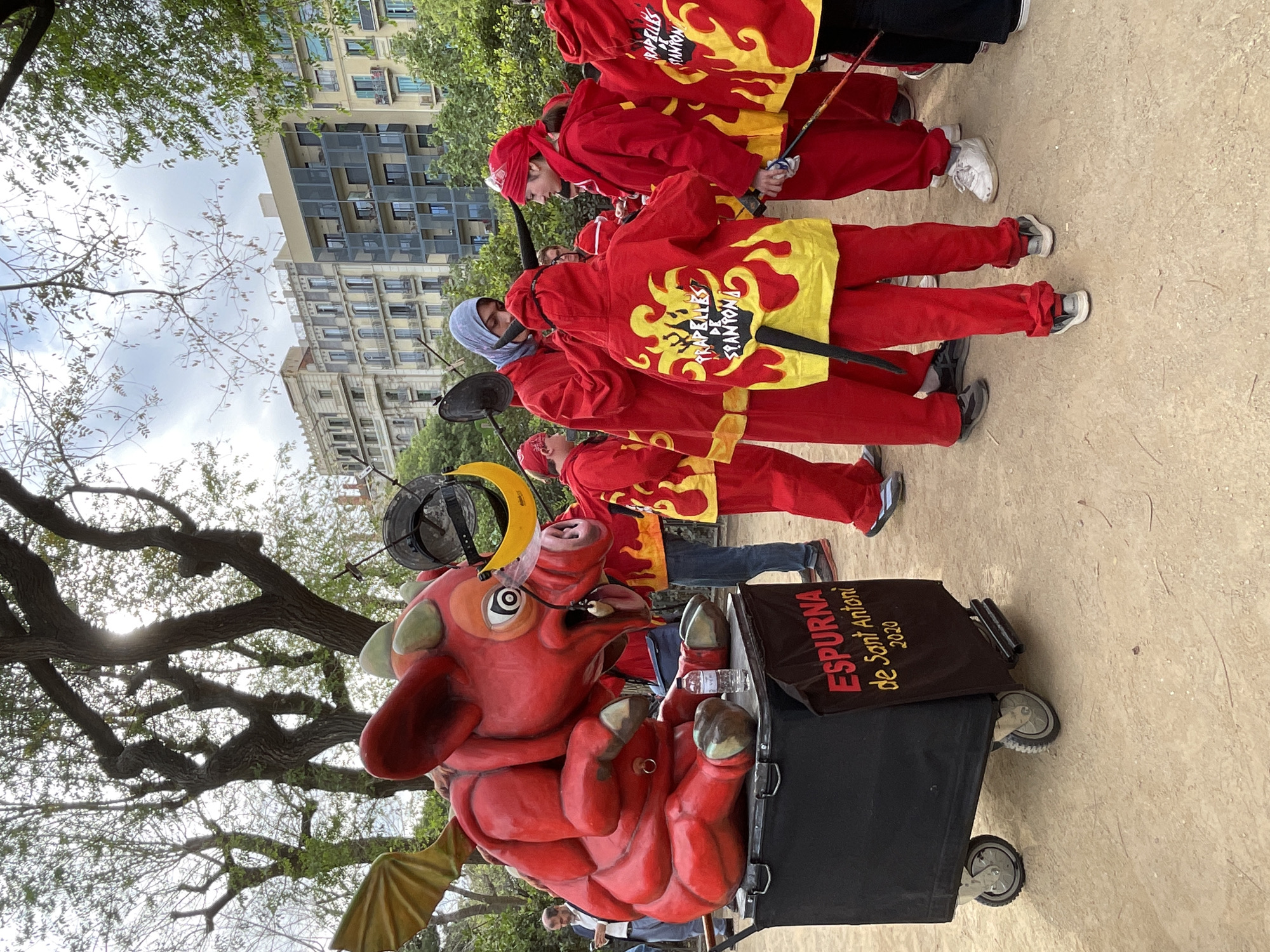 This daytime image captures a vibrant outdoor cultural procession, likely in Barcelona, Spain. The central focus is a group of individuals dressed in matching red and yellow robes, adorned with striking yellow flame patterns. Some participants wear head coverings, and several carry staffs or sticks, suggesting their role in a performance or parade. On the back of their costumes, the text "PAPALLELES ST. ANTONI" is visible in a flame-like font, likely indicating the name of their group and its association with Saint Anthony.
In the foreground, a large, elaborate red figure resembling a stylized pig or dragon with small wings is prominently displayed on a black wheeled cart. This "bestiari" figure features a prominent snout, large eyes, and metallic elements around its head. A black banner attached to the cart prominently displays the text "ESPURNA de Sant Antoni 2020." "Espurna," meaning "spark" in Catalan, likely refers to the figure's name, connecting it to fire-related festivities such as "Correfocs." The "Sant Antoni" reference points to the traditional feast day of Saint Anthony, and "2020" may indicate its creation year or a specific event. A plastic water bottle rests on the cart next to the figure.
The setting includes a dirt or gravel ground, with large trees featuring thick, gnarled branches and green foliage overhead, casting dappled light. In the background, various multi-story urban buildings are visible against a bright, overcast sky, characteristic of a city environment. The scene portrays a lively community event, showcasing traditional Catalan festival elements with participants in distinctive attire and an iconic "gegant" or "bestiari" figure.