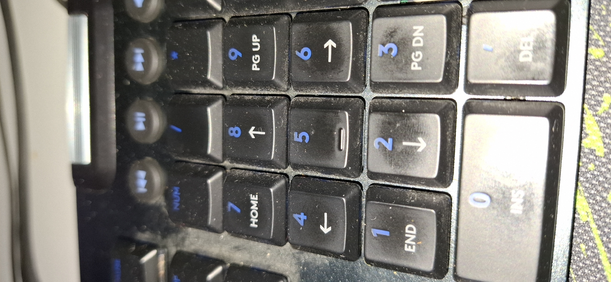 The image shows a close-up, top-down view of a black keyboard, focusing on the numeric keypad and some function keys. The keyboard appears to be slightly dusty. The numeric keys are arranged in a standard layout.
From left to right and top to bottom, the visible keys include:
* A row of keys with media control icons (rewind, play/pause, fast-forward) and "NUM" label.
* The number 7 key with "HOME" below it.
* The number 8 key with an upward arrow below it.
* The number 9 key with "PG UP" below it.
* The number 4 key with a leftward arrow below it.
* The number 5 key with a horizontal line below it.
* The number 6 key with a rightward arrow below it.
* The number 1 key with "END" below it.
* The number 2 key with a downward arrow below it.
* The number 3 key with "PG DN" below it.
* The number 0 key with "INS" below it.
* The decimal separator key (labeled with "/") and "DEL" key.
The keyboard is placed on a surface that has a green and black speckled pattern. The background is blurred and appears to be part of a desk or computer setup. There are no people or animals visible in the image. The lighting suggests an indoor environment. The image is taken at an angle that emphasizes the keys.