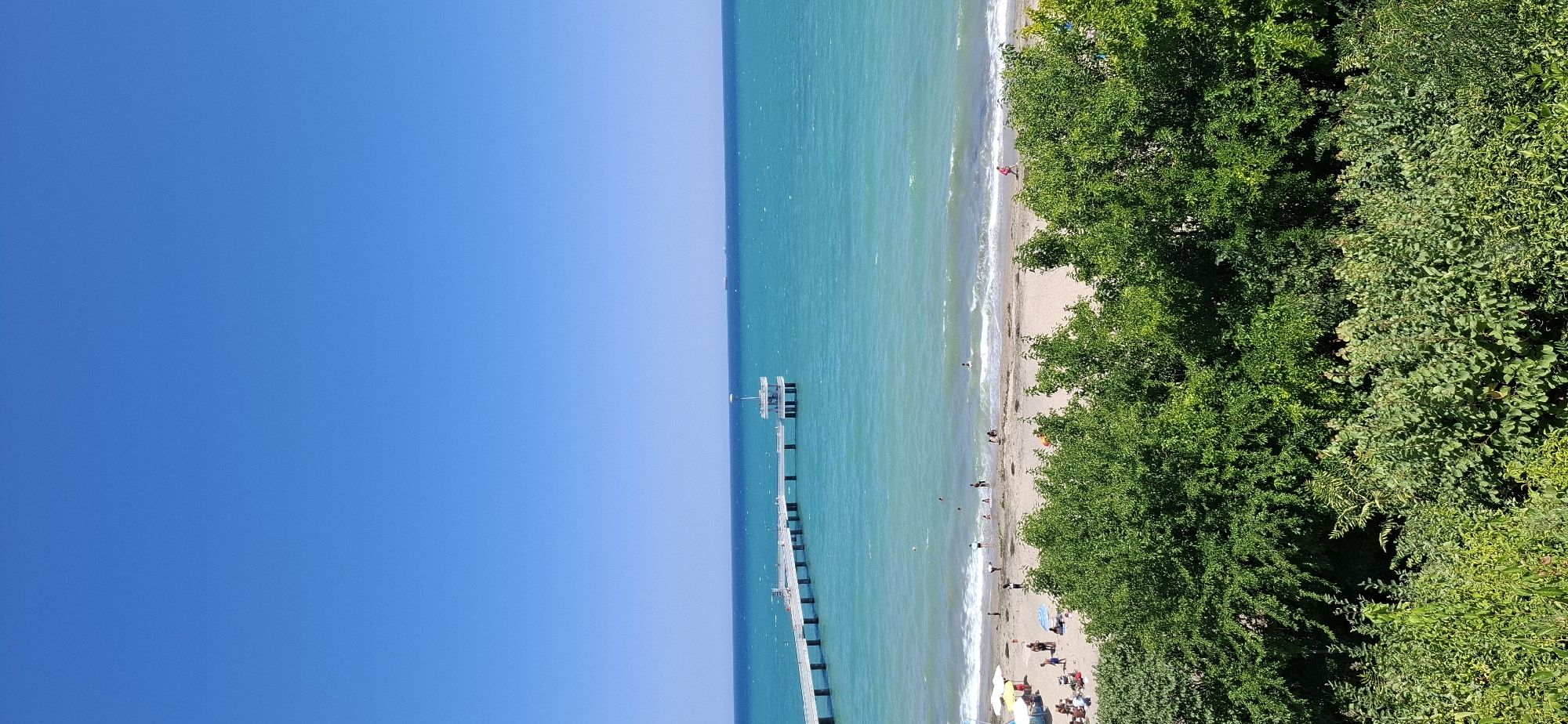 This is a photograph of the Burgas Sea Garden and the Burgas Pier. The image is taken from an elevated viewpoint looking out over the Black Sea on a clear, sunny day. The foreground is dominated by lush green trees and bushes. Beyond the foliage, a sandy beach is visible with several people sunbathing, walking, and wading in the shallow water. The turquoise sea stretches to the horizon, with small waves breaking on the shore. A long white pier extends out into the water, featuring a distinctive white, multi-level structure at its end. A few small boats or ships are visible in the distance on the horizon. The sky is a vibrant, cloudless blue. The scene depicts a typical summer day at the beach in Burgas.