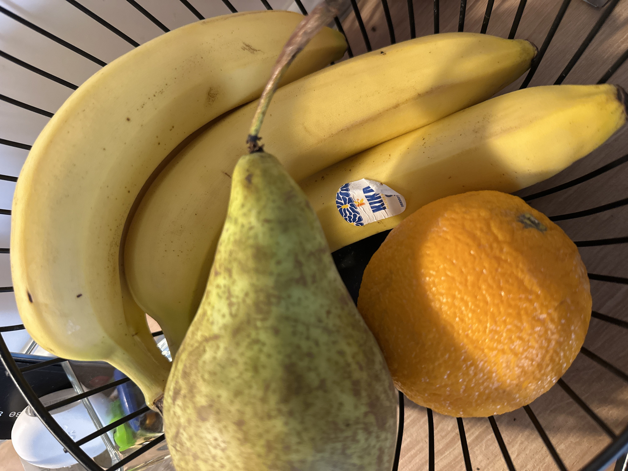 A high-angle shot shows a wire fruit basket containing three bananas, a pear, and an orange. The bananas are yellow and arranged at the top of the frame. The pear, positioned in the foreground, is greenish-yellow with brown speckles and has a stem. An orange is located on the right side of the basket. A small produce sticker is visible on one of the bananas. The setting is indoors, likely in Donji Pobori, Montenegro.