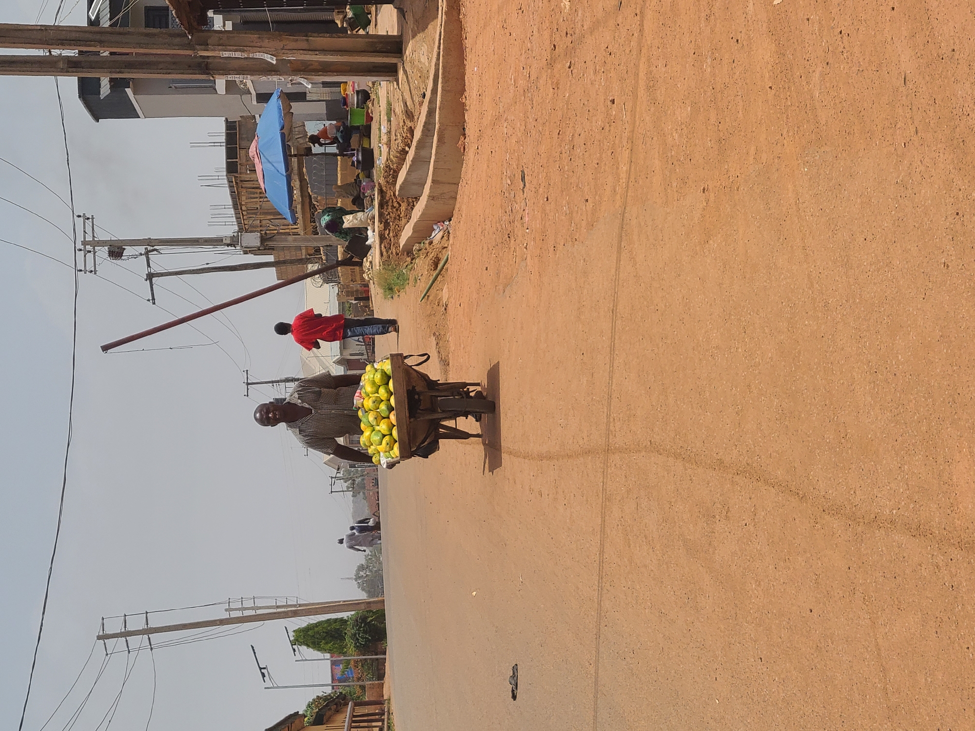A man pulls a small cart loaded with mangoes along a dirt road in Jos, Nigeria. The man is dark-skinned and wears a patterned shirt. The mangoes are green and yellow. A man in a red shirt stands on the side of the road near construction. Buildings, utility poles, and other pedestrians are visible in the background. The road is dusty. The sky is overcast.