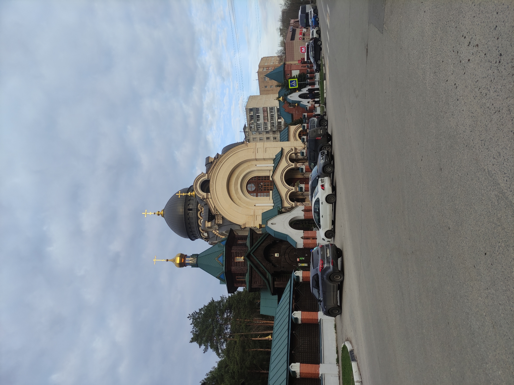 A street view presents a large church complex situated on the left side of the frame, with several parked vehicles lined up along the right side of the street. The church complex features a prominent structure with a dark dome and golden cross, adjoined by a smaller wooden building with a green roof and a golden-topped spire. Cars of varying colors, including white and gray, are parked neatly in a row along the curb. In the background, several buildings, including a light-colored apartment complex and other structures with varying heights and architectural styles, are visible. The sky is overcast with a mix of gray and blue hues. The location is in the city of Unknown, Unknown.