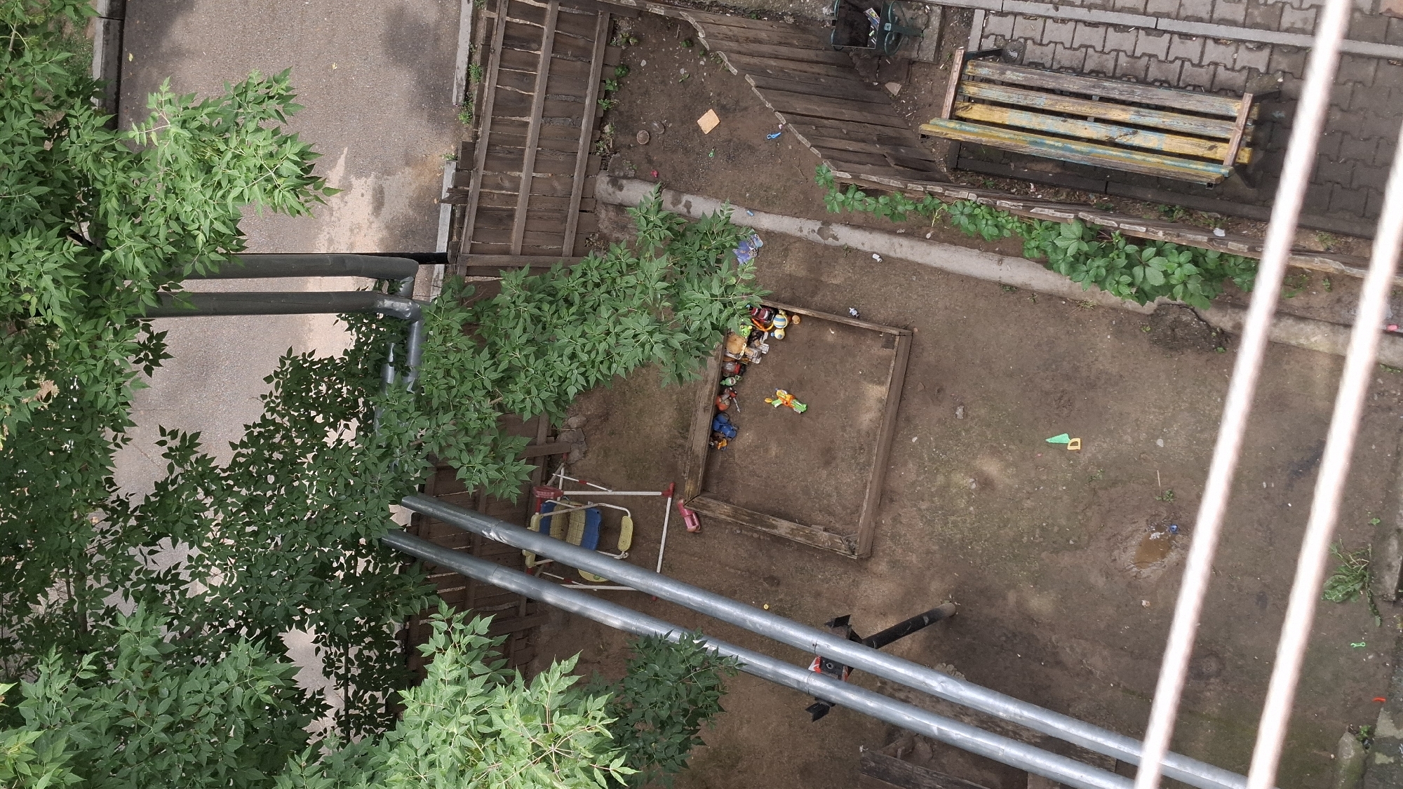 This image depicts an outdoor children's play area, likely located in a residential courtyard in Shymkent, Kazakhstan. The scene is viewed from an elevated perspective, possibly a balcony or window. The ground is a mixture of dirt and some paved sections. Prominently featured is a wooden sandbox filled with sand and various toys, including toy cars and figures. Adjacent to the sandbox is a swing set with a blue and yellow seat. Surrounding the play area is a wooden fence. In the background, a paved pathway and a wooden bench are visible. Trees with green foliage provide shade and greenery to the environment. The overall impression is one of a slightly unkempt but functional recreational space for children.