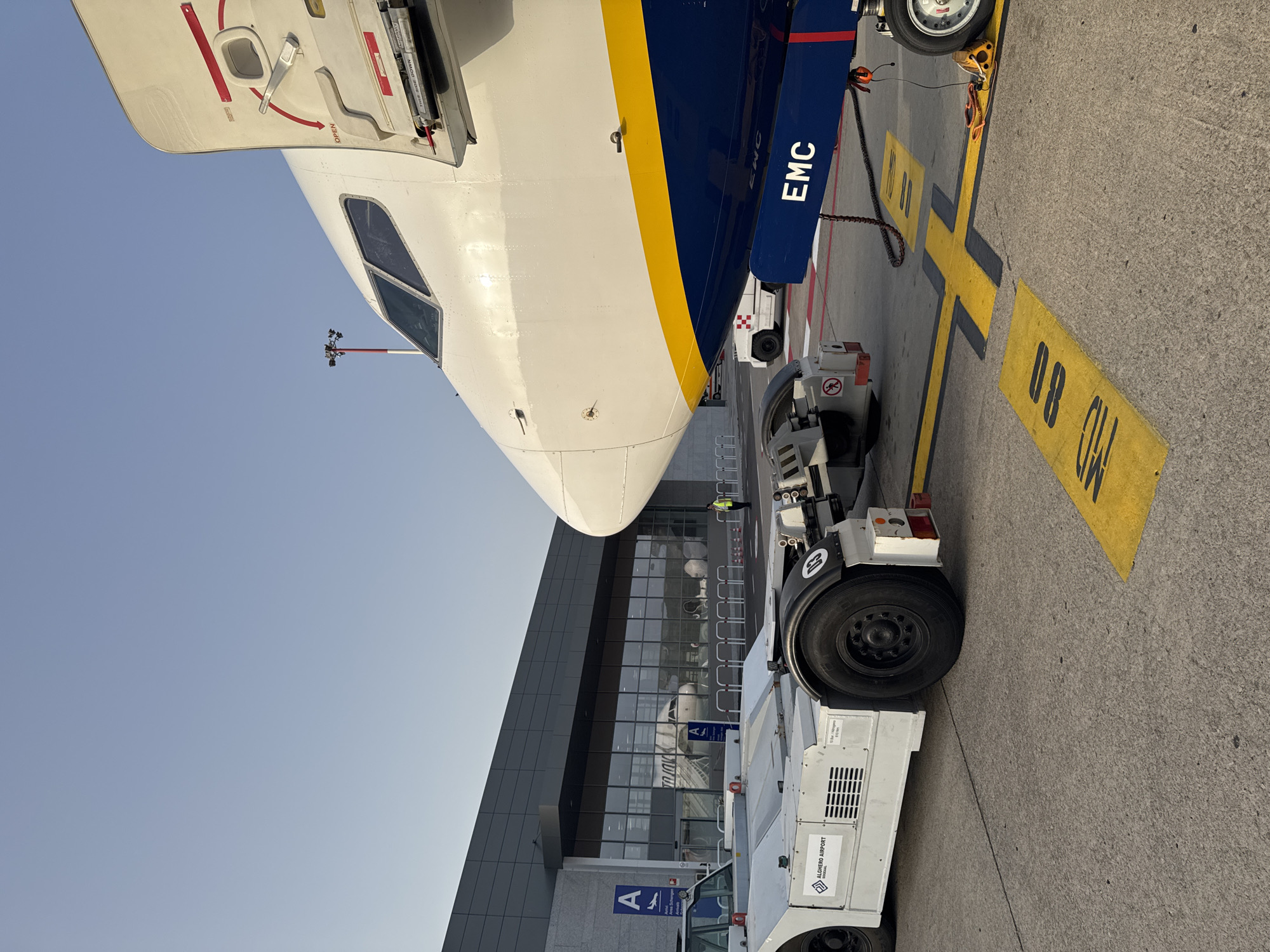 This is a close-up, ground-level, eye-level shot taken at Alghero Airport in Alghero, Italy. The image features the nose and forward fuselage of a white airplane with a blue and yellow livery. The aircraft's door is open, revealing the interior structure. In the foreground, a white airport tow tractor is positioned to the left of the aircraft. The tractor has "ALGHERO AIRPORT" printed on its side. A yellow and black painted line marking for the aircraft is visible on the tarmac in front of the plane. In the background, the modern terminal building of Alghero Airport is seen, with large glass windows reflecting the sky and aircraft. A lone worker in a yellow vest stands near the terminal entrance. The sky is clear and blue, indicating a bright, sunny day. The overall scene depicts an aircraft being serviced or prepared for departure at an airport.