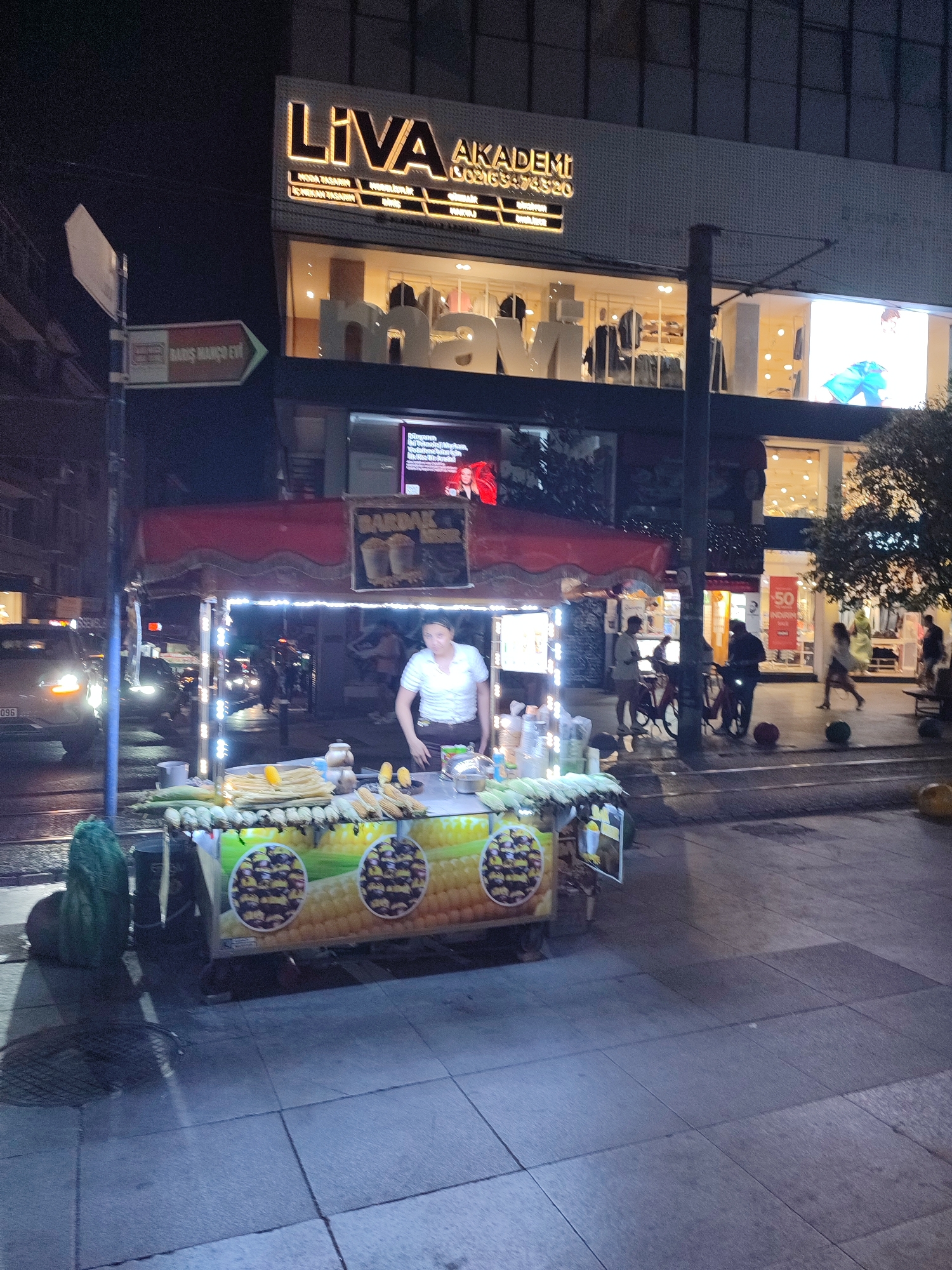 In Kadıköy, Turkey, at night, a street vendor is selling corn from a cart illuminated by string lights. The vendor, wearing a white shirt, stands behind the cart which displays rows of cooked corn on the cob and some raw corn. The cart is adorned with circular graphics of corn. In the background, a building with illuminated signage for "LIVA AKADEMİ" is visible. Pedestrians and vehicles are present on the street, with traffic lights and shop windows also illuminated. The scene is set on a paved sidewalk next to a street with tram tracks.