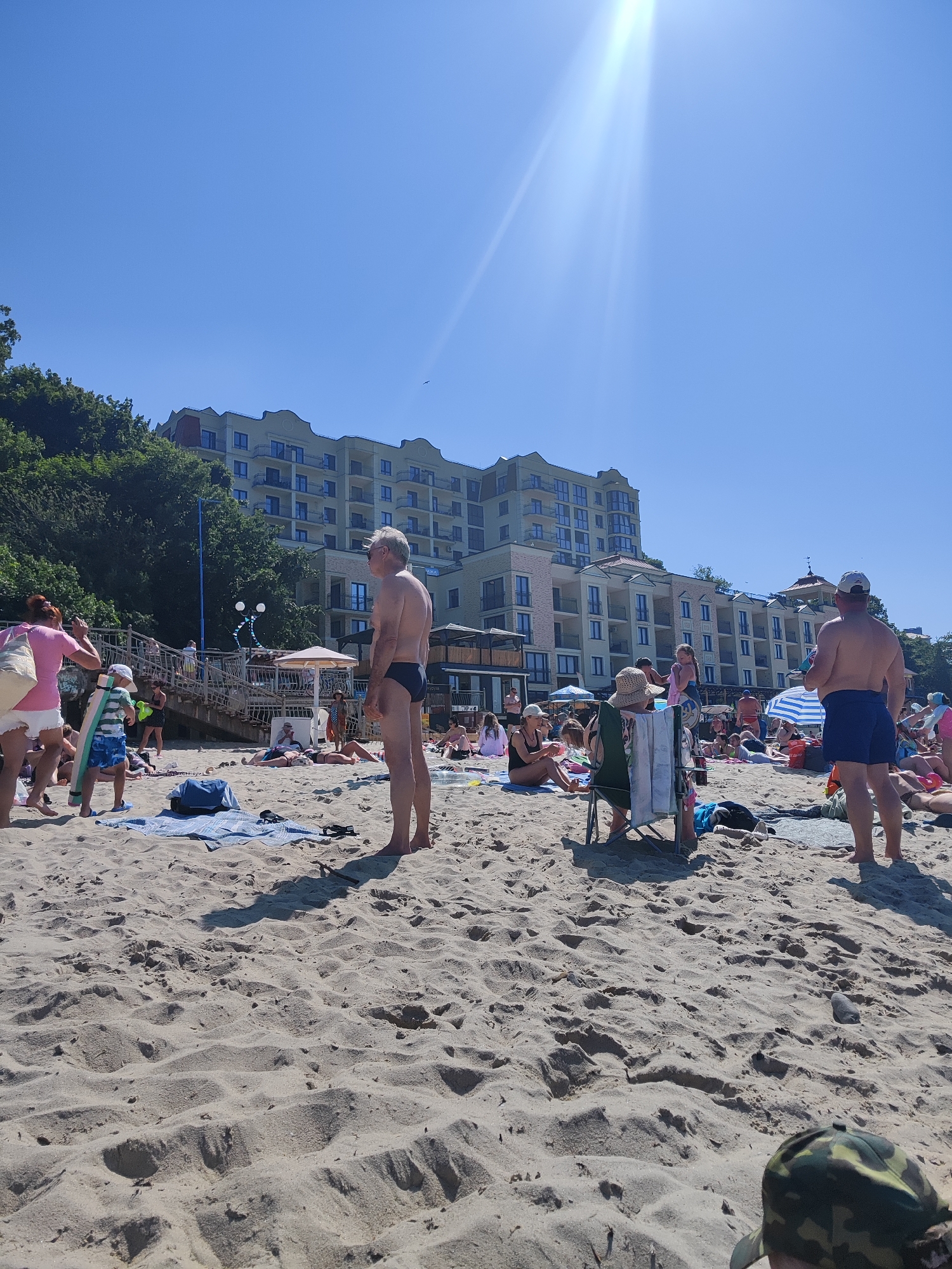 This is a photograph taken on a sunny day on the beach in Pionersky, Russia. The beach is sandy and appears to be crowded with people. In the background, there is a large, modern, multi-story hotel or apartment building situated on a hill covered with trees. The sky is clear and blue with bright sunlight streaming down, creating lens flares.
On the beach, there are many people in various states of relaxation and activity. Some are sunbathing on towels and blankets, while others are standing or walking. A few people are partially obscured by other beachgoers or objects.
In the foreground, a man in dark swimming trunks stands facing away from the camera. To his left, another man, also in dark swimming trunks, is captured in profile, looking towards the right side of the frame. A young boy wearing a striped shirt and blue patterned shorts is holding a boogie board and is positioned to the left of the man in profile. Nearby, a woman in a pink t-shirt and white shorts is also present.
Several beach umbrellas and chairs are scattered across the sand. There are also bags and towels visible on the beach. The overall scene depicts a typical summer day at a popular beach resort.