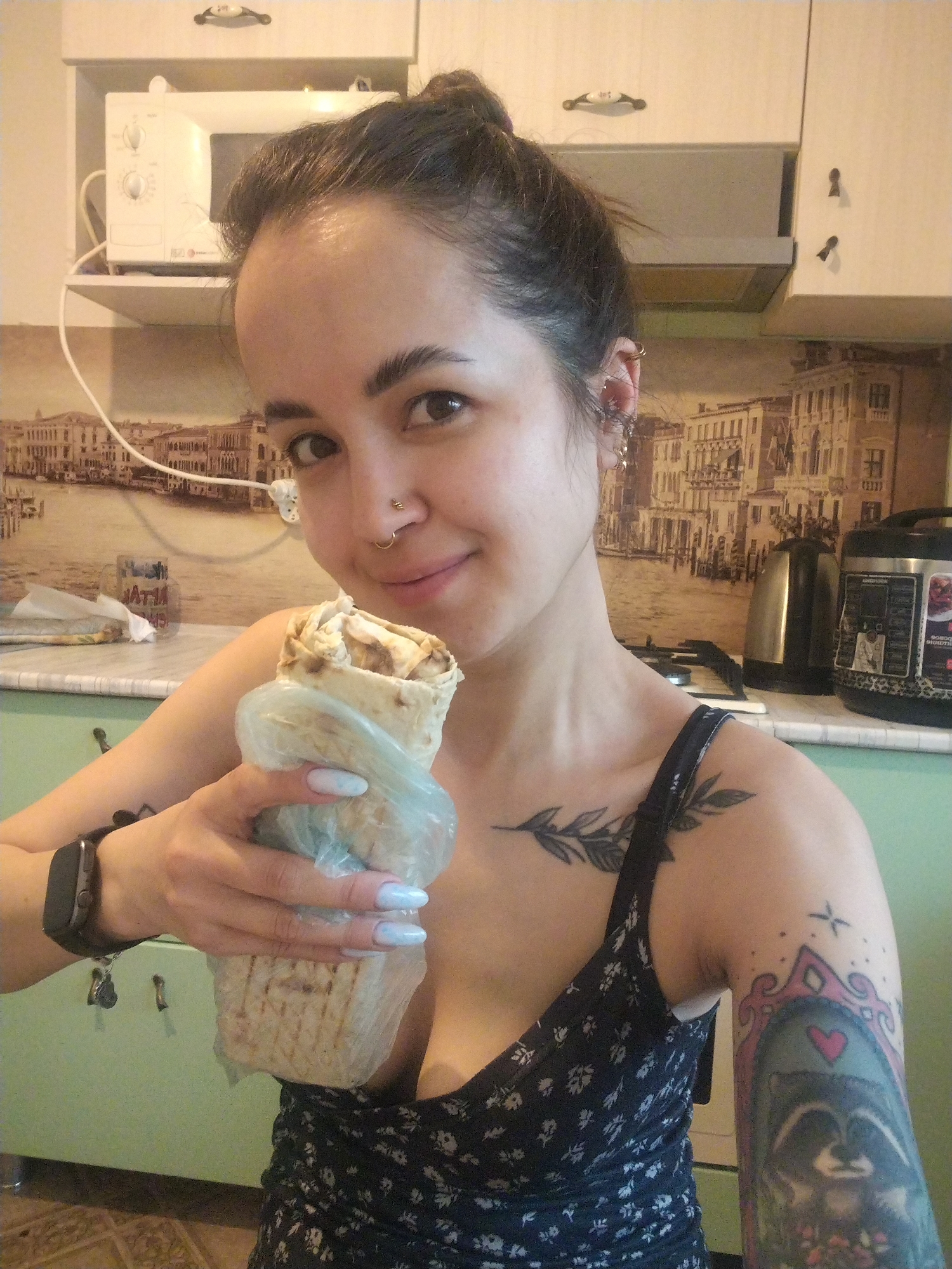 A woman holds a shawarma in a kitchen. She has dark hair pulled into a bun, fair skin, a nose ring, and multiple ear piercings. She is wearing a black floral-patterned top. She has a leafy branch tattoo on her chest, and a raccoon tattoo on her right arm. She holds a shawarma wrapped in clear plastic in her left hand. She has a smartwatch on her left wrist. The kitchen has light green walls and cabinets. There is a microwave on a shelf above the counter, and a backsplash depicting a cityscape. An electric kettle and a multi-cooker are on the counter. Keys are hanging from a cabinet handle. The city is not specified and information about Noviy, Russia is not discernible from the image.