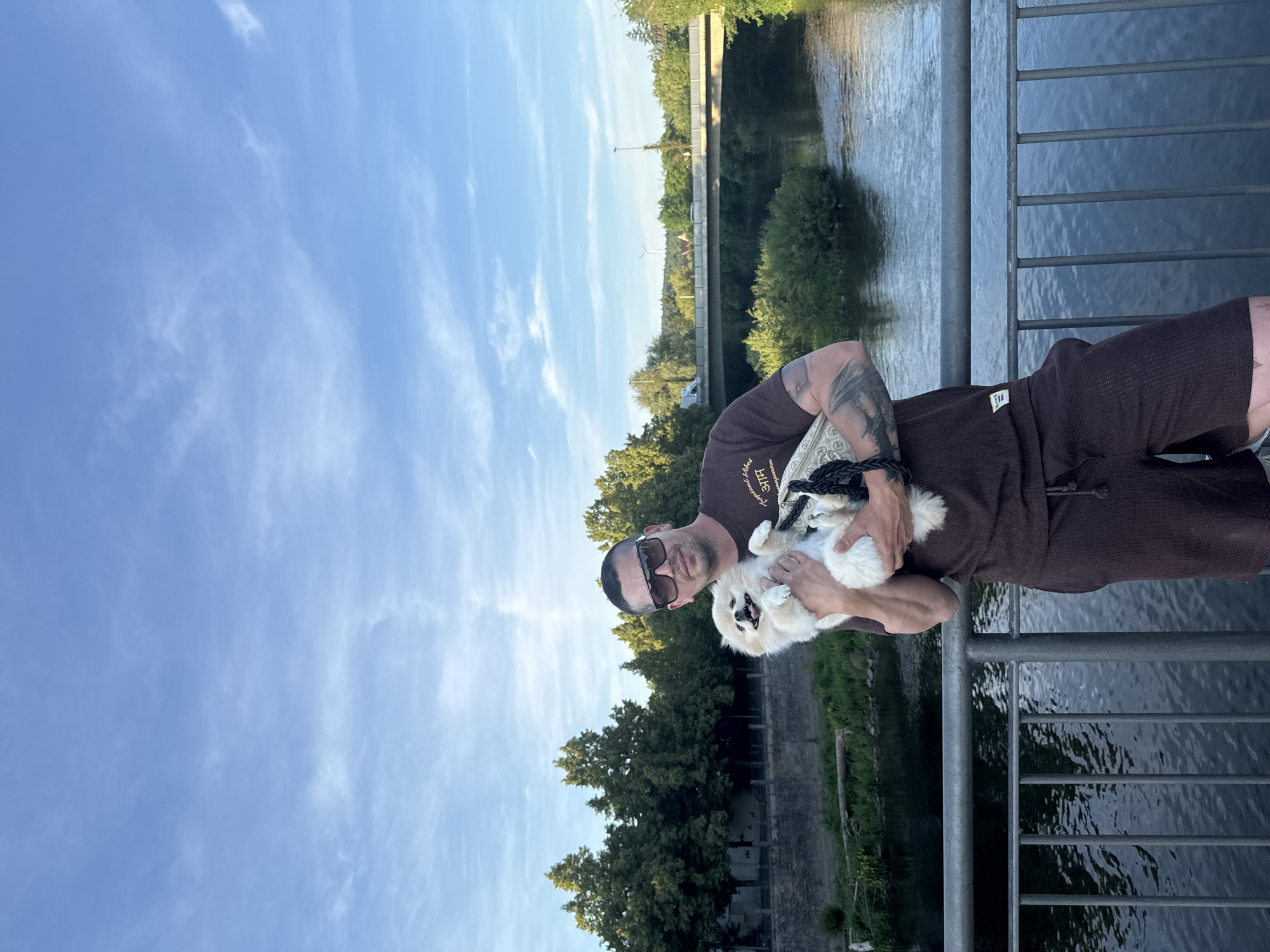 A man is holding a white Pomeranian dog while standing on a bridge in Bietigheim-Bissingen, Germany. The man is wearing sunglasses, a brown t-shirt, and brown shorts. He has tattoos on his left arm. The bridge has metal railings, and a river flows underneath. The sky is blue with wispy clouds. Trees line the riverbank.