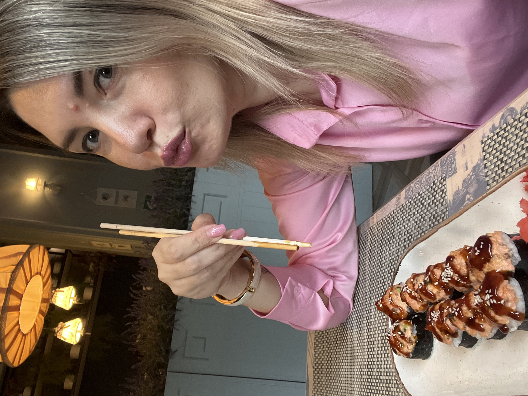 A woman with blonde hair and pink lipstick is holding chopsticks and looking at the camera with her lips pursed. She is wearing a pink silk shirt and a gold bracelet. In front of her is a plate of sushi rolls. The scene appears to be in a restaurant with hanging lanterns and lavender plants. The location is the city of Sochi, Russia.