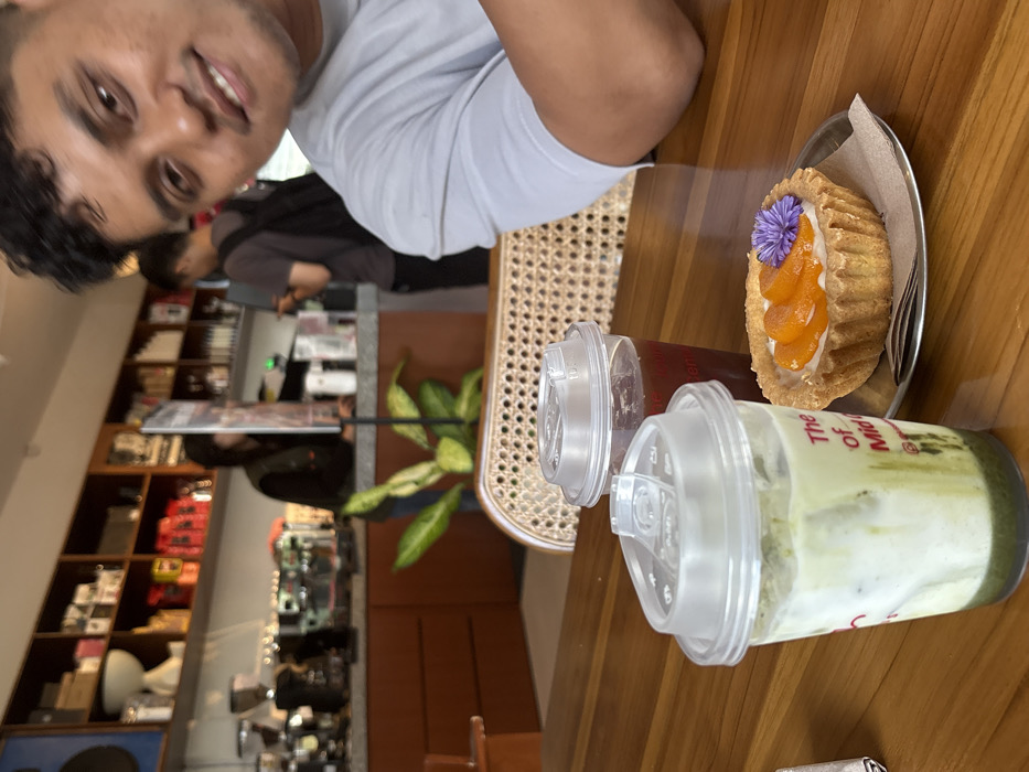 An indoor scene in a cafe in Kloncing, Indonesia, depicts a close-up perspective of a table with beverages and a pastry, with a man in the foreground and the cafe interior in the background.
In the immediate foreground, to the right, a male individual with dark hair and a light gray t-shirt is visible from the chest up, looking directly at the camera with a slight smile. On a wooden table to his left, two clear plastic cups with clear domed lids are present. The cup closest to the viewer contains a layered green and white liquid, consistent with an iced matcha latte. The second cup, positioned behind the first, contains a dark reddish-brown liquid, likely iced coffee or tea. Adjacent to these drinks, a single round tart with a fluted crust, topped with slices of orange fruit (possibly apricots or peaches) and a small purple flower, rests on a small, circular metallic plate with a piece of brown parchment paper underneath. A wicker-backed chair is partially visible behind the table, between the foreground and background.
The background shows the interior of a cafe. A long service counter with a reddish-brown base and a light gray top extends across the frame. Behind the counter, various espresso machines and coffee-making equipment are visible. Shelving units line the wall behind the counter, displaying numerous items including coffee bags, cups, and bottles. Two individuals are present in the background: one person, partially obscured and wearing dark clothing, stands behind the counter, while another person wearing a dark t-shirt and a backpack stands facing the counter, possibly placing an order. A large potted plant with variegated green and yellow leaves is positioned on the floor in front of the counter. The overall lighting is bright and even.