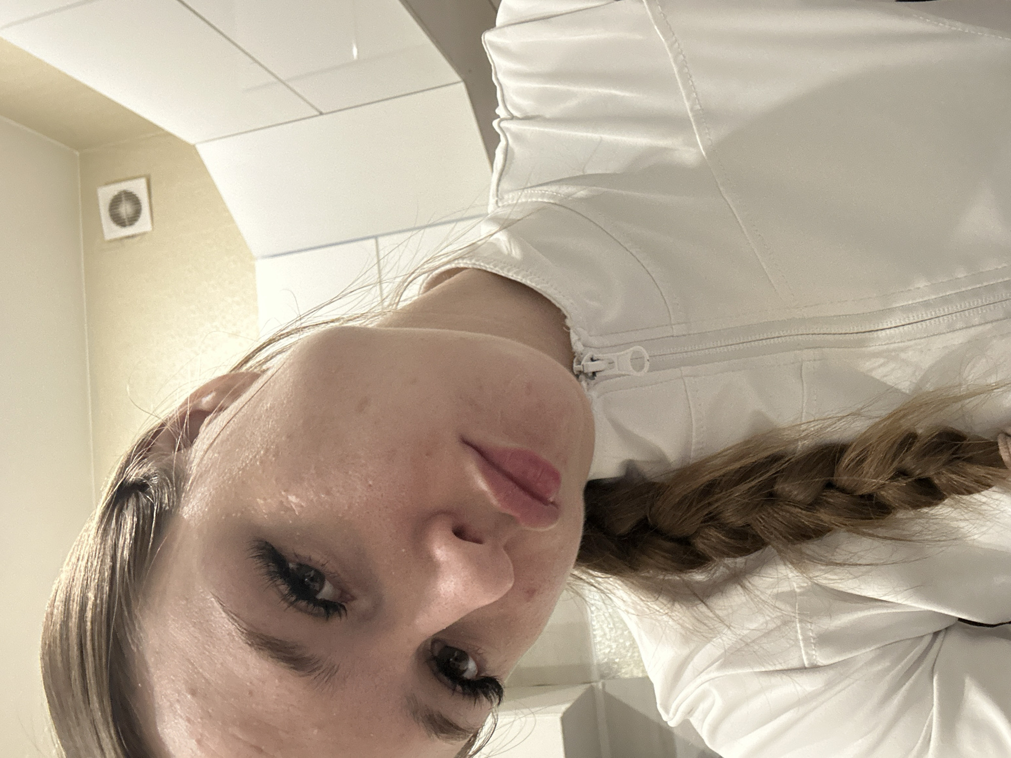 A close-up, slightly angled shot shows a person with light skin and brown hair braided down their back. They are wearing a white, zippered jacket. The background suggests a bathroom setting with white tiles and a ventilation fan on the wall. The location in the city of Unknown, Unknown is not specified.
