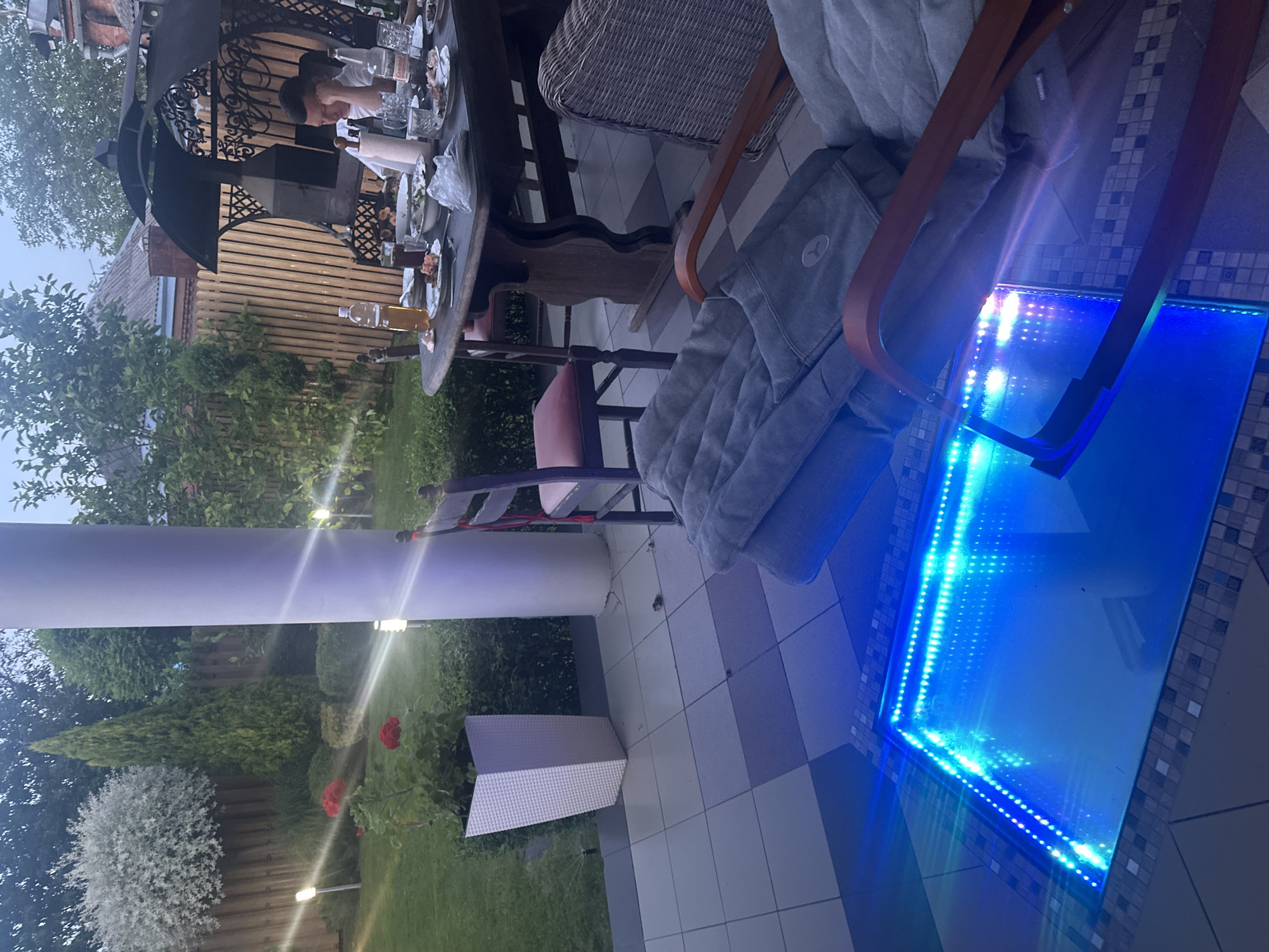 The image depicts an outdoor patio area, likely in Armavir, Russia, during twilight. A partially consumed meal is laid out on a rustic wooden table, suggesting a recent gathering. One person, a young male wearing a white t-shirt, is seated at the table, looking down with his hand supporting his head, possibly appearing tired or contemplative. The table is adorned with plates, glasses, bottles, and food remnants.
Surrounding the table are several chairs, including a dark wooden chair with a red cushion and a wicker chair. In the foreground, a modern seating arrangement features a chair covered with gray cushions, hinting at a comfortable lounging area.
The environment showcases a well-maintained garden with manicured bushes and trees. Two white outdoor lamps illuminate the lawn. A tall, white cylindrical pillar stands prominently on the patio, near a large white planter containing red roses. A unique feature of the patio is a rectangular recessed area filled with glowing blue LED lights, creating a striking visual effect. The flooring is tiled in a checkerboard pattern of white and blue-grey squares. In the background, a wooden fence encloses the property, and glimpses of a building with a reddish-brown roof can be seen. The overall atmosphere is one of relaxed evening leisure.