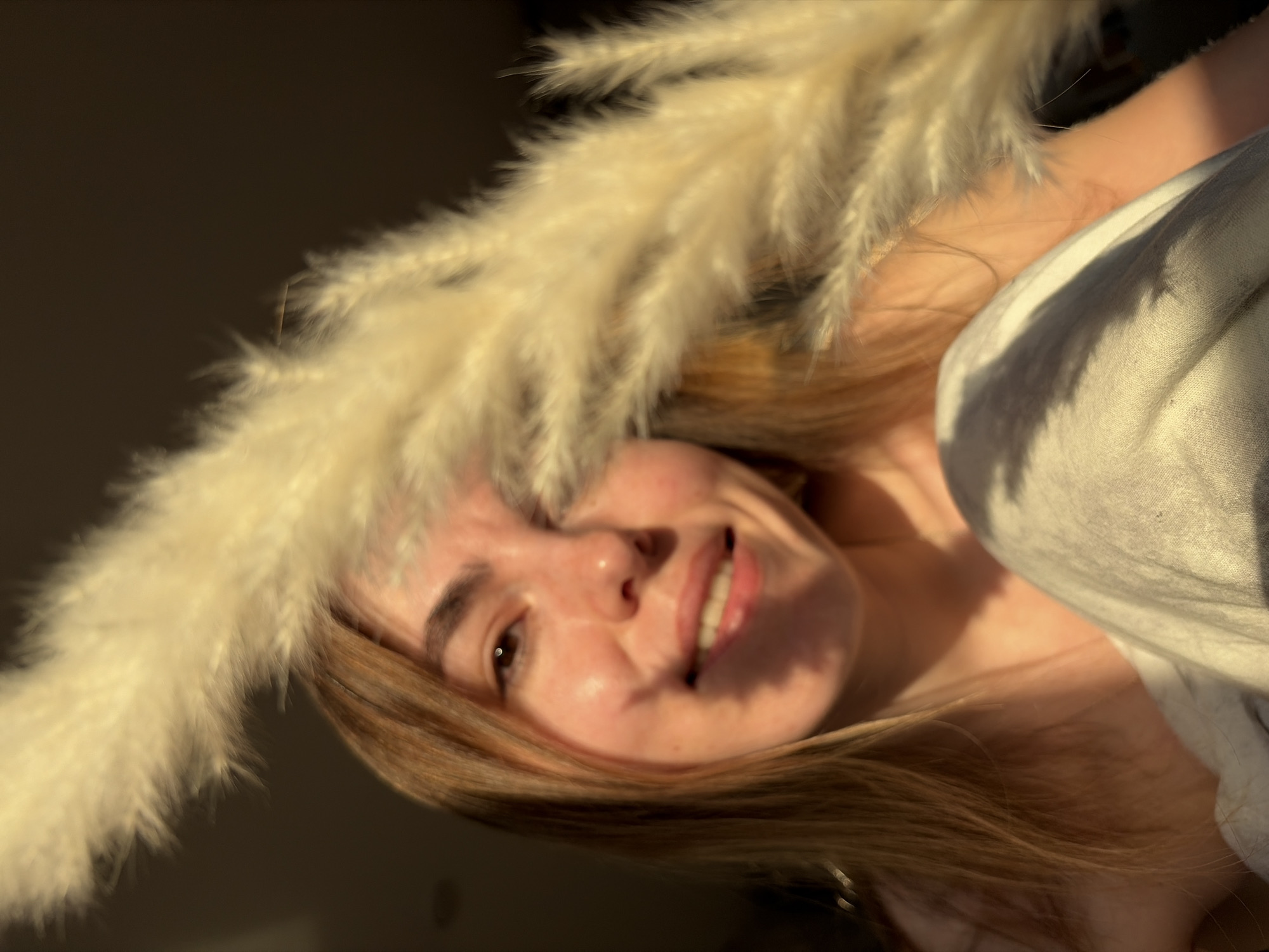 A woman is smiling at the camera while holding dried pampas grass near her face. She is wearing a white shirt or covering. The lighting is soft and warm, suggesting late afternoon or early morning. The location cannot be determined from the image.