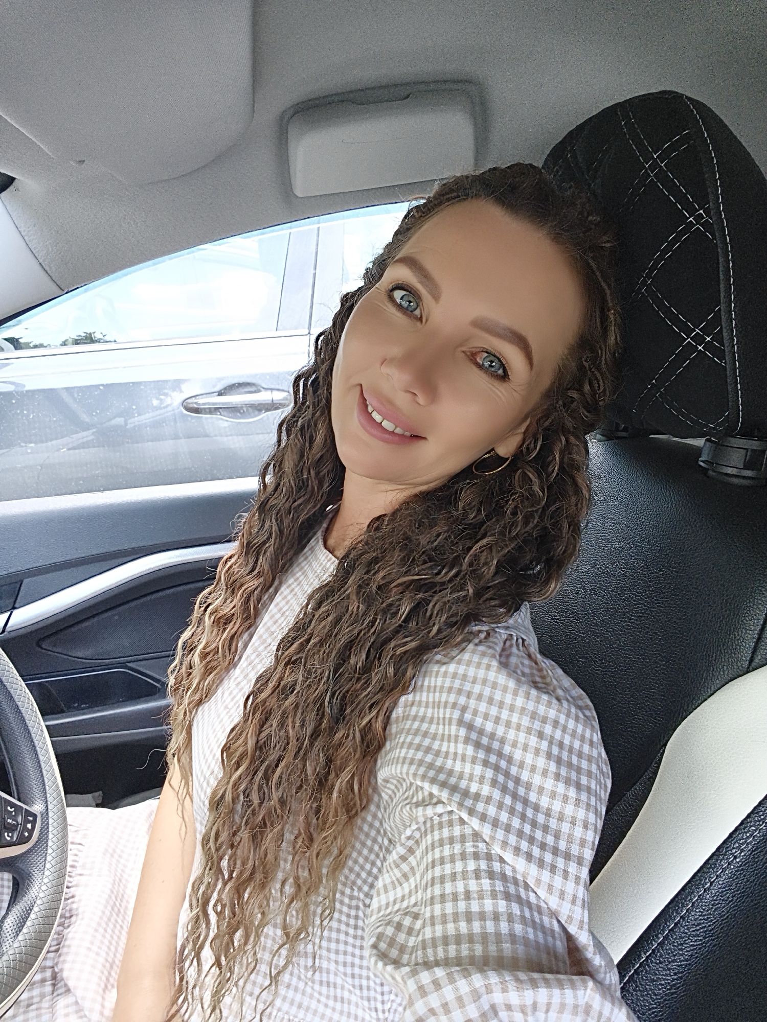 A woman with long, curly brown hair and blue eyes smiles at the camera from the driver's seat of a car. She is wearing a light-colored, checkered dress. The interior of the car is visible, including the steering wheel, dashboard, and part of the passenger seat. Outside the car window, another car is parked. The scene appears to be outdoors, likely in a parking lot.