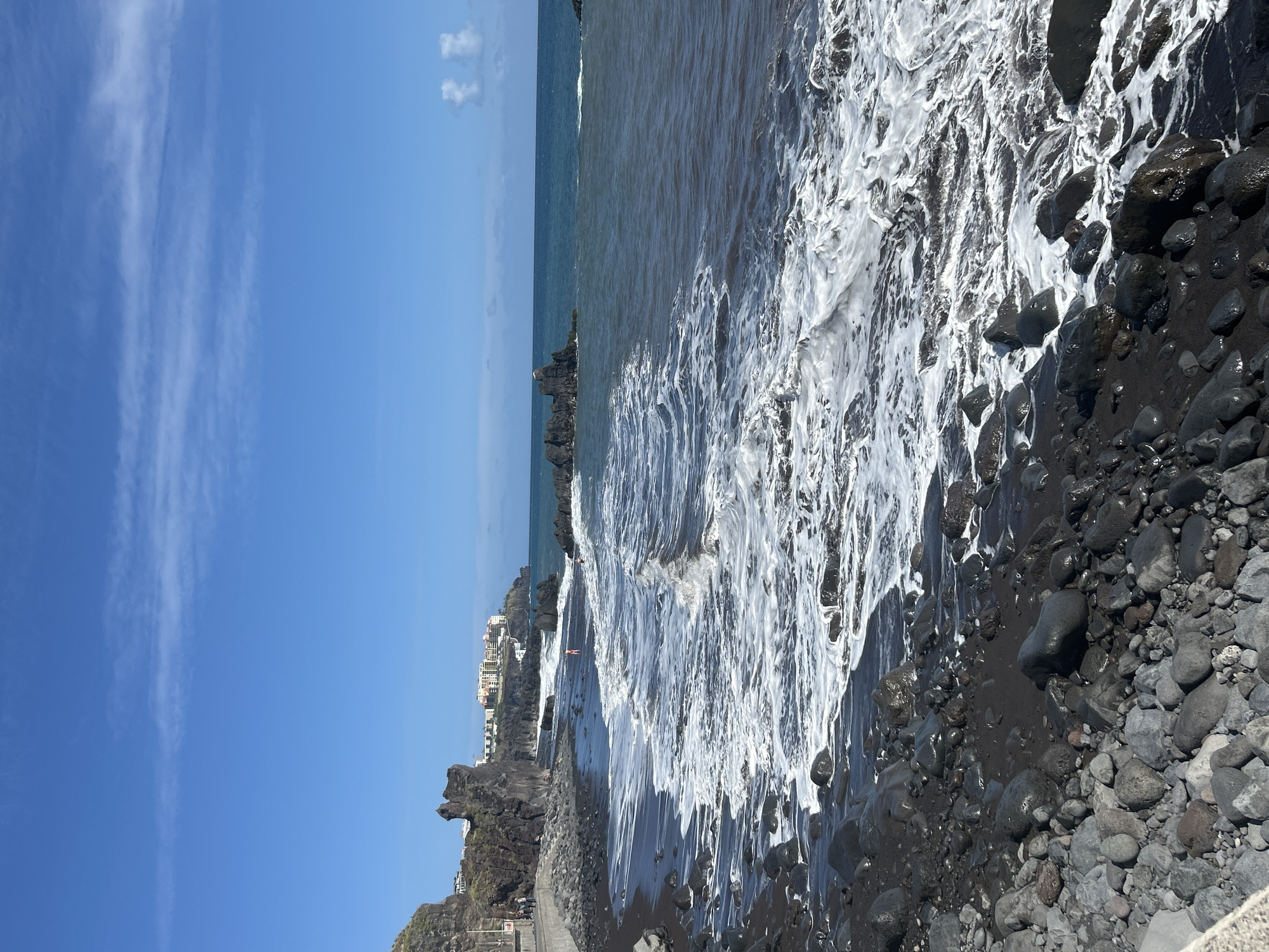 The image shows a black volcanic rock beach in Funchal, Portugal, with waves crashing against the shore. A clear blue sky with few clouds is overhead. The water is a dark grey, with white foam from the breaking waves dominating the scene along the shoreline. Dark grey and black rocks and pebbles cover the beach. Buildings and rocky outcroppings are visible along the coastline in the background. A small number of people can be seen in the water.