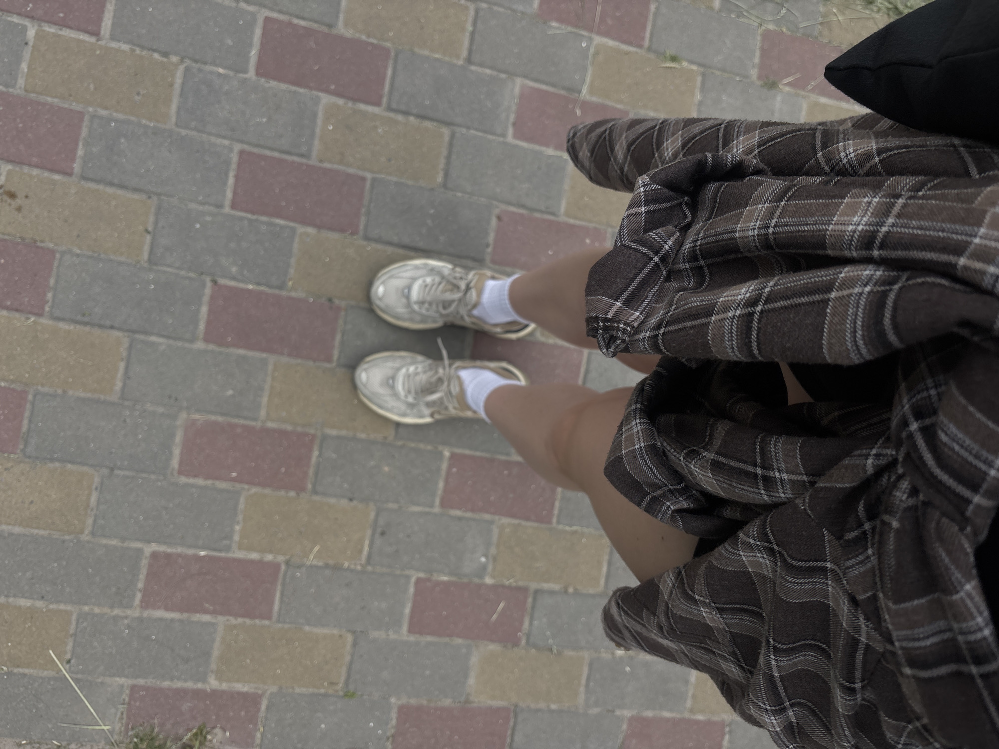 The image shows a close-up, overhead view of a person's legs and feet standing on a paved walkway. The walkway is made of rectangular bricks in varying shades of gray, red, and yellow. The person is wearing white socks and light-colored sneakers. A plaid skirt, in shades of brown and white, covers part of the person's legs and is draped over the edge of the frame. A black object, possibly a bag, is visible in the upper right corner. The scene appears to be outdoors, given the paving stones and a hint of grass or weeds in the lower left and right corners. The lighting suggests it is daytime, but the weather is unclear. No text is visible in the image. The focus is on the textures of the clothing and the walkway, creating a somewhat abstract composition.