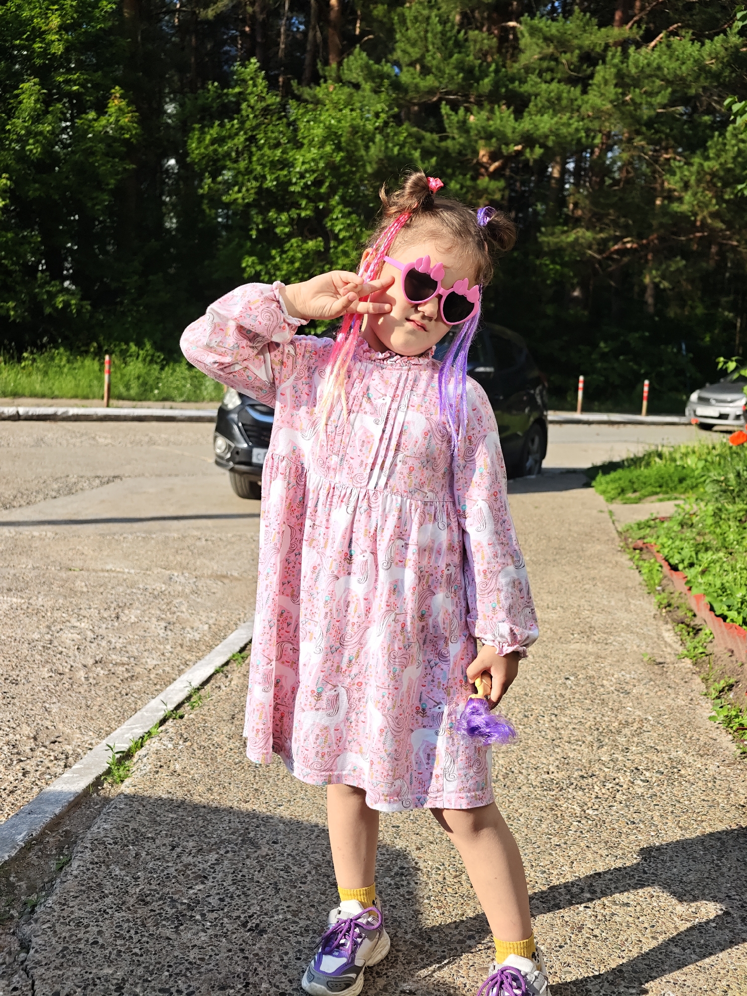 A young girl with pink and purple hair extensions, wearing heart-shaped pink sunglasses, a pink unicorn-print dress, yellow socks, and white and purple sneakers, stands on a paved walkway outdoors. She is holding a purple toy. Behind her, a dark-colored car is parked on an asphalt surface, with a grassy area and trees in the background. The setting appears to be a residential or public area in Chaikovsky, Russia, given the typical architecture and vegetation.