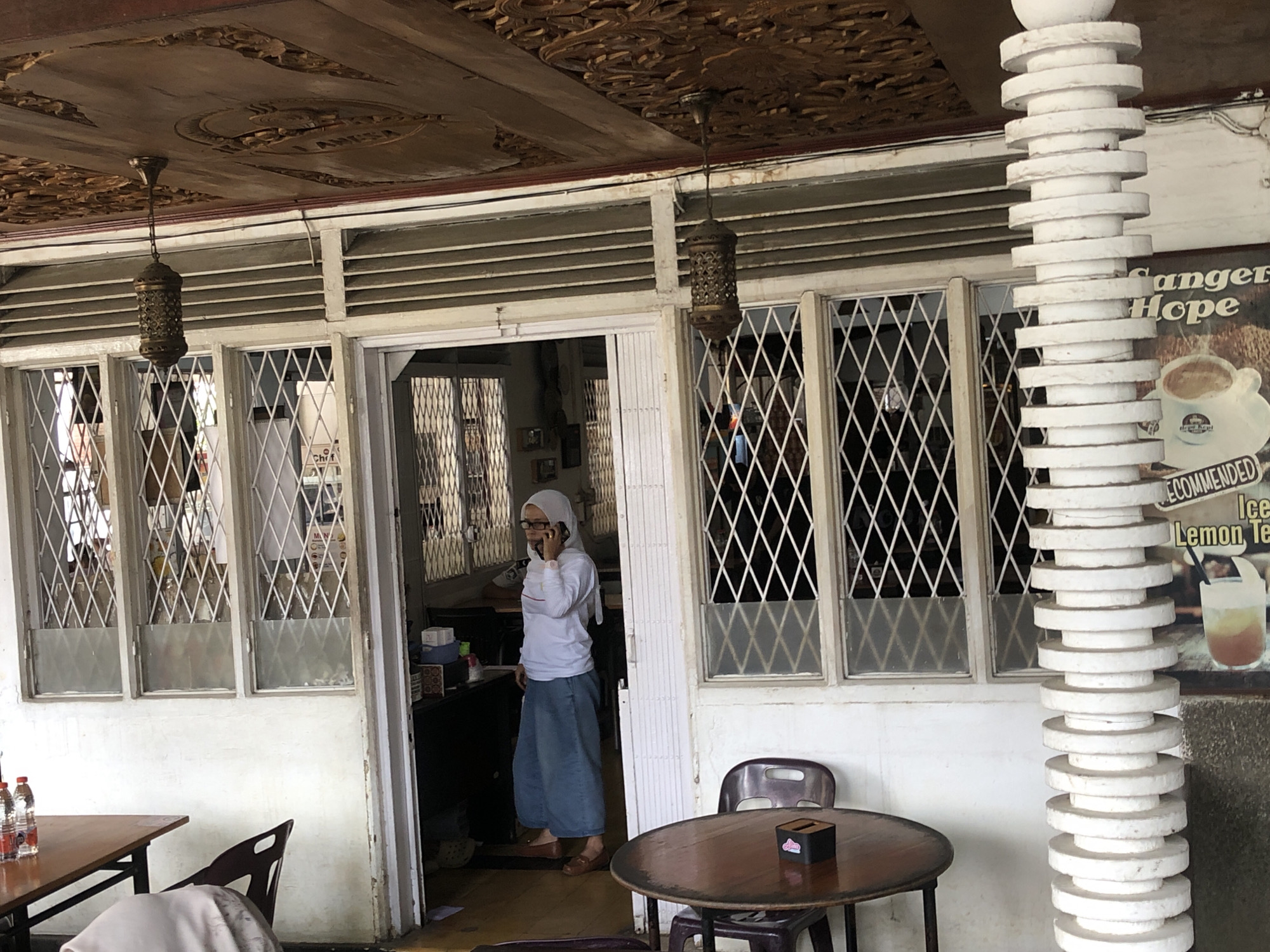 A woman wearing a white long-sleeved shirt, blue pants, and a white hijab is standing in a doorway, talking on a cellphone. The woman is wearing glasses and brown shoes. She is standing in front of a building with an open doorway. Inside the building, there are shelves and boxes. The building has windows with diamond-shaped grilles. Outside the building, there is a wooden table with two bottles of water on it, and a dark chair. To the right of the doorway, there is a round wooden table with a tissue box on it, and a dark chair. There is also a white cylindrical pillar with stacked circular elements to the right of the building. A sign with text that includes "Sanger", "Hope", "Recommended", "Ice", and "Lemon Tea" is visible behind the pillar. The ceiling of the building is adorned with intricate carvings. The overall scene appears to be an outdoor seating area of a cafe or restaurant in Langsa, Indonesia.