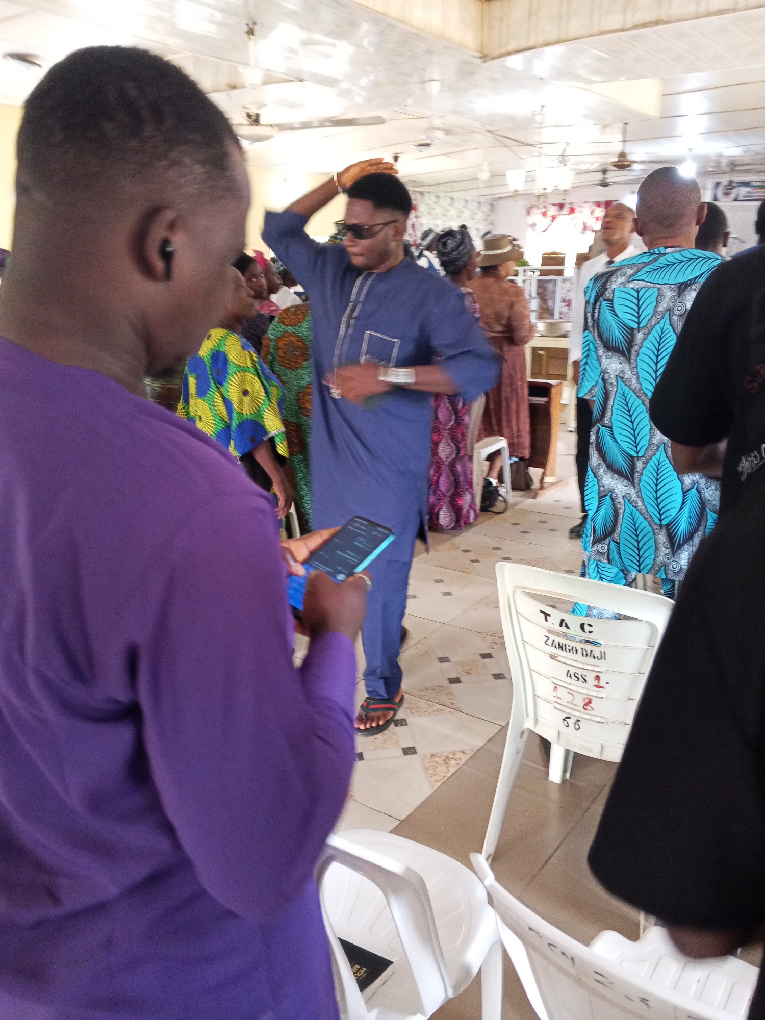 In Zango-ndaji, Nigeria, a group of people are gathered indoors, likely for an event, as indicated by the festive attire and seating arrangements. The room appears to be a hall or community center with a high ceiling, featuring fluorescent lighting and ceiling fans. The floor is tiled with a geometric pattern.
In the foreground, a person wearing a vibrant purple outfit is turned away from the camera, holding a smartphone in their left hand. Their attention is directed downwards towards the screen. To their right, a man in a blue kaftan is standing, with his right arm raised as if gesturing or adjusting something. He is wearing sunglasses and appears to be interacting with others in the vicinity.
Further back, other individuals are visible. One woman is wearing a patterned headwrap and a brown dress, while another is adorned in a colorful, patterned fabric. A man in the background sports a striking blue and black patterned kaftan, facing away from the camera.
Several white plastic chairs are arranged throughout the space. One chair in the middle ground has text written on its backrest, including "T. A. C," "ZANGO DAJI," "ASS 1," and numbers that are partially visible. This suggests the event may be associated with a specific organization or an assigned seating arrangement.
The overall atmosphere appears to be social and possibly celebratory, with individuals engaged in conversation or observing the proceedings. The presence of personal devices and the attire suggest a modern gathering within a Nigerian cultural context.