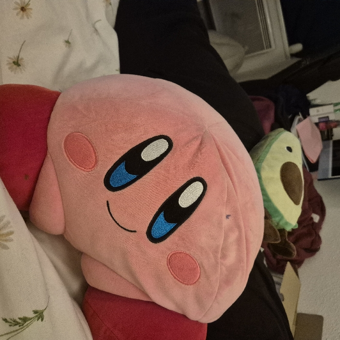 The scene presents an interior view of what appears to be a bedroom, located in Strasbourg, France. In the immediate foreground, a large, plush pink Kirby toy is prominently displayed, resting on a white bedsheet patterned with small, light-colored floral designs. The Kirby toy faces slightly to the right, exhibiting its signature black and blue eyes, small pink blush circles on its cheeks, and a subtle black curved line forming a smile. Red fabric, consistent with Kirby's feet, is visible on either side of its spherical body. Behind the Kirby plush, partially obscured by dark fabric, a green avocado-shaped plush toy with a brown central pit is visible. The background is primarily composed of dark, indistinct fabric or clothing, with a white, horizontal radiator or heating unit visible on the far left. No individuals are present in the frame.