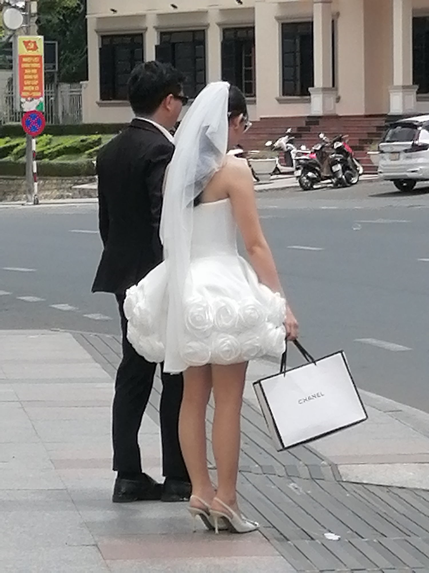 In Da Lat, Vietnam, a man in a black suit and sunglasses stands on a sidewalk, facing away from the camera. Beside him, a woman in a white wedding dress with a veil also faces away. Her dress features a voluminous skirt adorned with rosettes. She carries a white Chanel shopping bag in her right hand. In the background, a white car is parked near a row of buildings and several motorcycles are parked on the street. A traffic sign with red and blue circular elements is visible on a pole to the left.