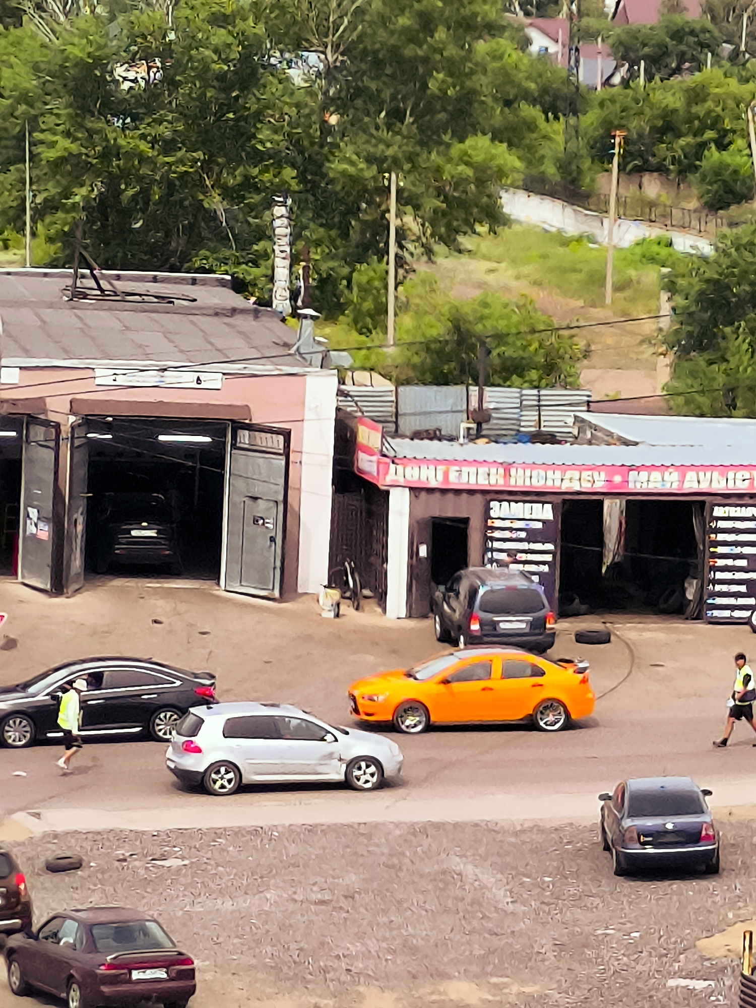 The image depicts an outdoor scene at a car repair shop in Qarağandı city administration, Kazakhstan. Several vehicles are present: a black sedan, a silver hatchback, an orange sedan, a dark SUV, and a dark purple coupe in the foreground. A person wearing a yellow vest and dark shorts is walking near the black sedan, and another person in a yellow vest and dark shorts is walking near the orange sedan. The car repair shop features multiple open bays, some with cars inside. Signs are visible above the bays, one with pink lettering and another with white text on black backgrounds. The environment includes a dirt/gravel lot in the foreground and a grassy, wooded hillside in the background with some buildings visible at the top. The weather appears to be overcast.