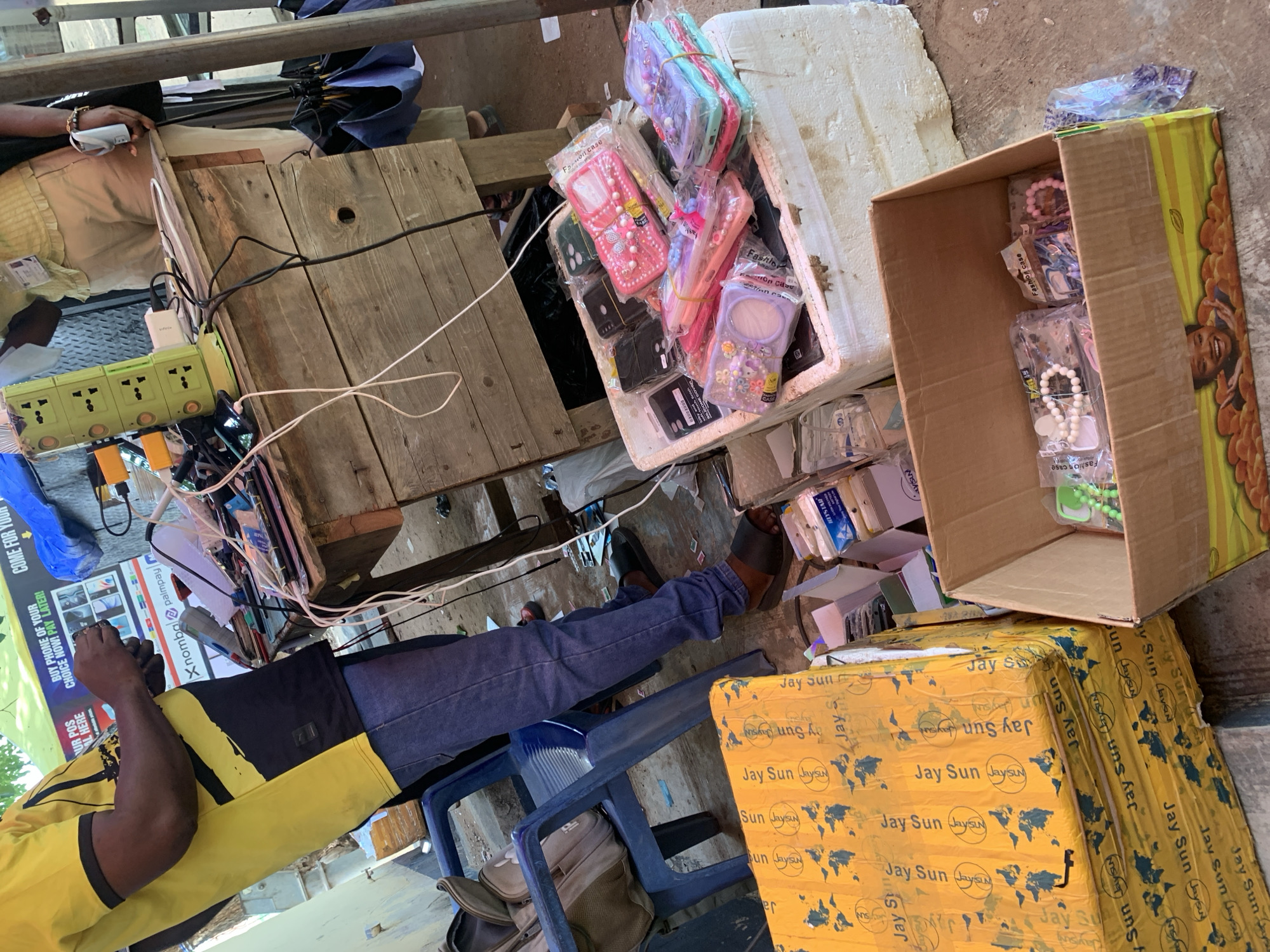 This image captures an informal marketplace or roadside stall, likely in Ilorin, Nigeria, specializing in phone accessories and possibly charging services. The scene is brightly lit, suggesting daytime.
In the foreground, a person wearing a yellow and black t-shirt and blue jeans is seated on a blue plastic chair, looking intently at a mobile phone in their hand. Another individual, dressed in light-colored clothing, is partially visible in the upper left, also seemingly engaged with a device or power bank.
The stall setup is informal, utilizing wooden planks as surfaces. A prominent green multi-socket power strip, with an "Infinix" branded charger and multiple cables plugged in, suggests phone charging is a key service or that multiple devices are being managed.
The primary merchandise appears to be phone cases. Numerous "Fashion case" phone covers, some described as "SILICON," are displayed in transparent plastic packaging. They come in various colors like pink, purple, and blue, with some featuring decorative elements such as pearls or charms. These are arranged in an open styrofoam box and a large cardboard box. Another large yellow cardboard box, repeatedly branded with "Jay Sun (Jaysun)" and a map graphic, serves as a stand for other items.
Visible text on a sign advertises, "BUY PHONE OF YOUR CHOICE NOW! PAY LATER!" and states, "OUR POS IS AVAILABLE HERE." The sign also mentions "Xnomba palmpay." Smaller labels like "NISA HYSTAR" are also discernible in the background. The overall impression is a busy, practical commercial setting.
