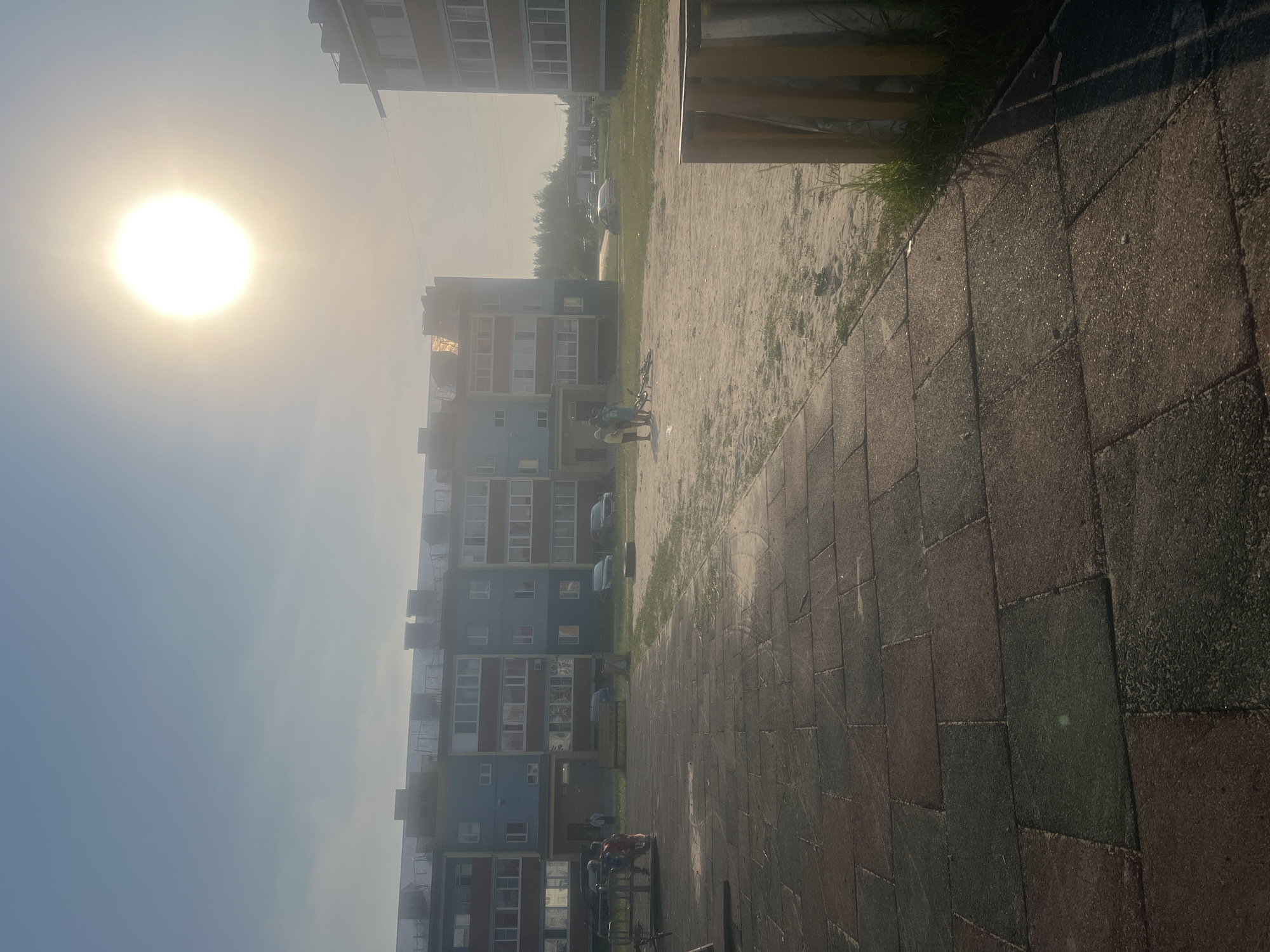 In a city environment, a sun-drenched scene unfolds. The foreground features a paved area with large, dark rectangular tiles. To the left, a section of a dark grey playground structure is visible. Beyond this, a sandy courtyard is present, with patches of dry grass. In the middle ground, a light blue apartment building with red trim and multiple balconies is the dominant structure. Several cars are parked in front of it, and a park bench sits near the entrance. To the right of this building, another apartment building, primarily red brick with white trim and balconies, is partially visible. In the courtyard, a group of four young people are gathered, with one appearing to be speaking. A bicycle lies on the ground nearby. In the background, a line of trees is visible against a hazy sky. The sun, a bright, luminous circle, is prominently positioned in the upper right portion of the frame, casting a strong glare and creating lens flare. The overall atmosphere is bright and sunny, suggesting a warm day.