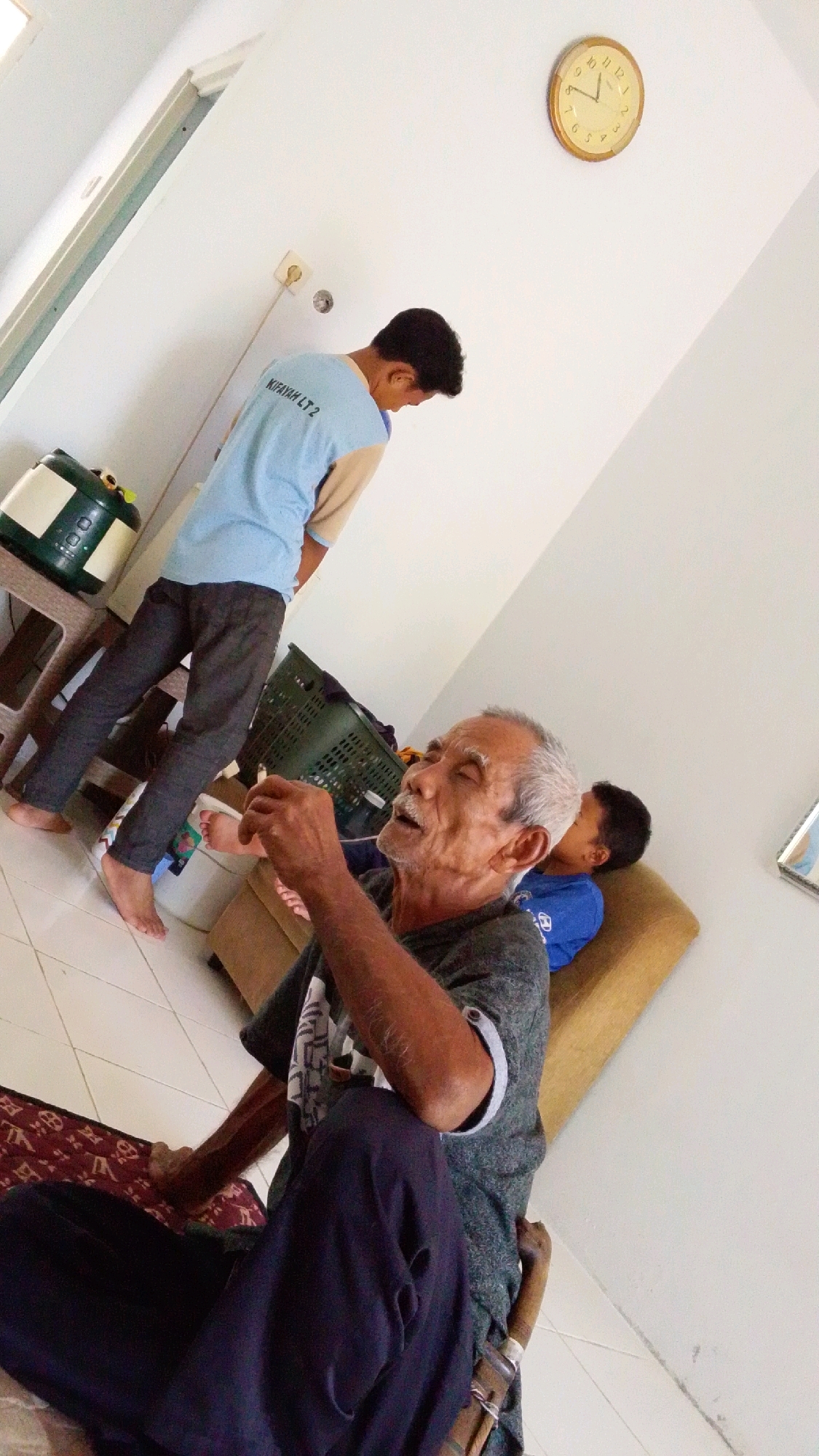An elderly man with gray hair and a mustache is seated on the floor, smoking and looking upwards. He is wearing a dark gray t-shirt and dark pants. Next to him, a young boy in a blue shirt is leaning against a couch. Another young man in a light blue and tan shirt is standing in the background, looking down. A green laundry basket is on the floor behind the elderly man. A clock on the wall shows the time as approximately 12:35. The scene appears to be indoors, in a room with white walls and tiled flooring.