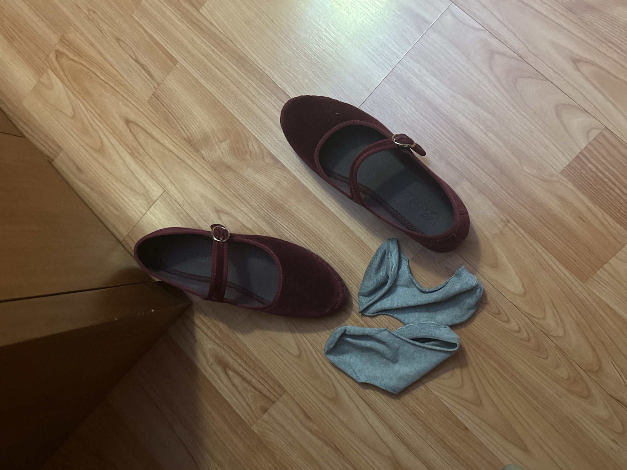 A pair of dark red Mary Jane shoes and a pair of light gray socks are on a light brown wood-grain floor. The location is not discernible from the image.