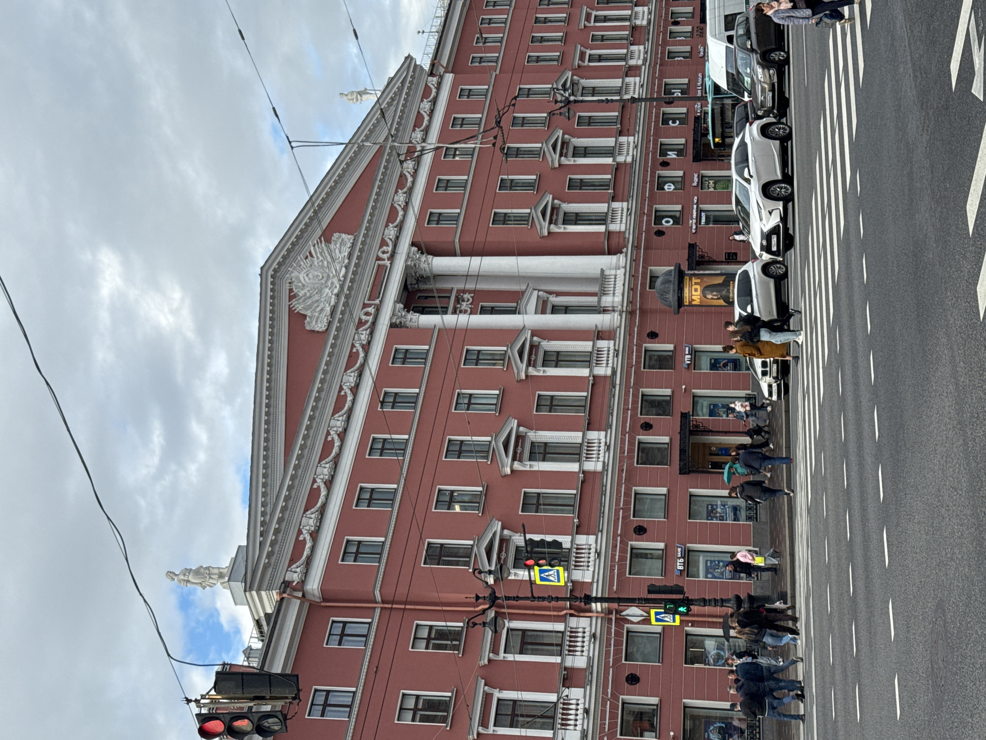 This is a vertical, eye-level, medium shot of a street scene in Saint Petersburg, Russia, on an overcast day. The foreground features a crosswalk on a wide asphalt road, with white dashed lines indicating lanes. Several people are crossing the street, some under umbrellas, suggesting it may be raining or has recently rained. Traffic lights are visible, with one showing red and another showing green.
Dominating the background is a large, ornate, red-brick building with many windows and decorative architectural elements, including columns and statues. The building appears to be several stories high. Cars are parked along the street and a few are in motion.
On the building's facade, there are signs with Russian text. One sign reads "ВТБ БАНК", indicating a VTB Bank. Another sign shows the word "MOT" in large letters, likely an advertisement. A billboard features a picture of a person's face.
The overall mood of the image is somewhat somber due to the cloudy weather, but the presence of people suggests normal urban activity. The architecture strongly suggests a European city, and the text confirms the location as Russia. The time of day is indeterminate due to the overcast conditions, but it appears to be daytime.