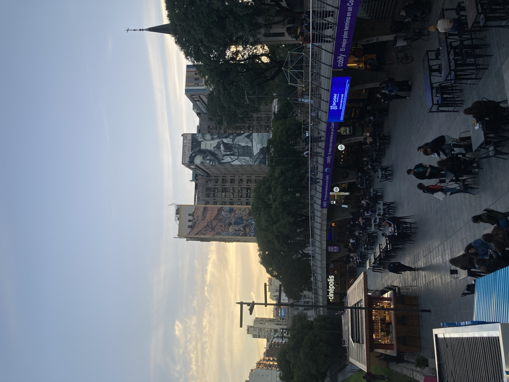 This image captures a vibrant urban scene in Buenos Aires, Argentina, during the late afternoon or early evening, characterized by the warm, golden light of the setting sun. The sky transitions from a clear light blue overhead to a soft yellowish-orange hue near the horizon.
The scene depicts a bustling pedestrian square or public space with numerous people socializing and dining at outdoor cafes and restaurants. Dark tables and chairs are densely arranged, indicating popular outdoor eating areas. Individuals are seen seated, chatting, and walking through the lively environment.
In the background, several multi-story buildings frame the area. A prominent building features a large, striking monochrome mural depicting various figures. Another significant structure clearly displays "HOSPITAL DE CLÍNICAS" on its facade, identifying it as the Hospital de Clínicas José de San Martín, a well-known landmark in Buenos Aires. A distant church spire topped with a cross is visible against the sky.
Throughout the scene, several digital screens and signs display advertisements primarily in Spanish. Notable visible text includes "cabify El mejor plan termina en un Cabif" (repeatedly for the ride-sharing service Cabify), "SanCorSalud La buena salud es todo" (for an Argentine health insurance company), and "cinépolis" (for the cinema chain). Other partial texts like "EQUUS" are also present.
Mature trees are interspersed among the buildings and public areas, adding greenery to the urban landscape. A modern kiosk or food stand is visible, alongside a metal pedestrian bridge. The overall impression is one of a lively, social urban gathering place under pleasant, clear skies.