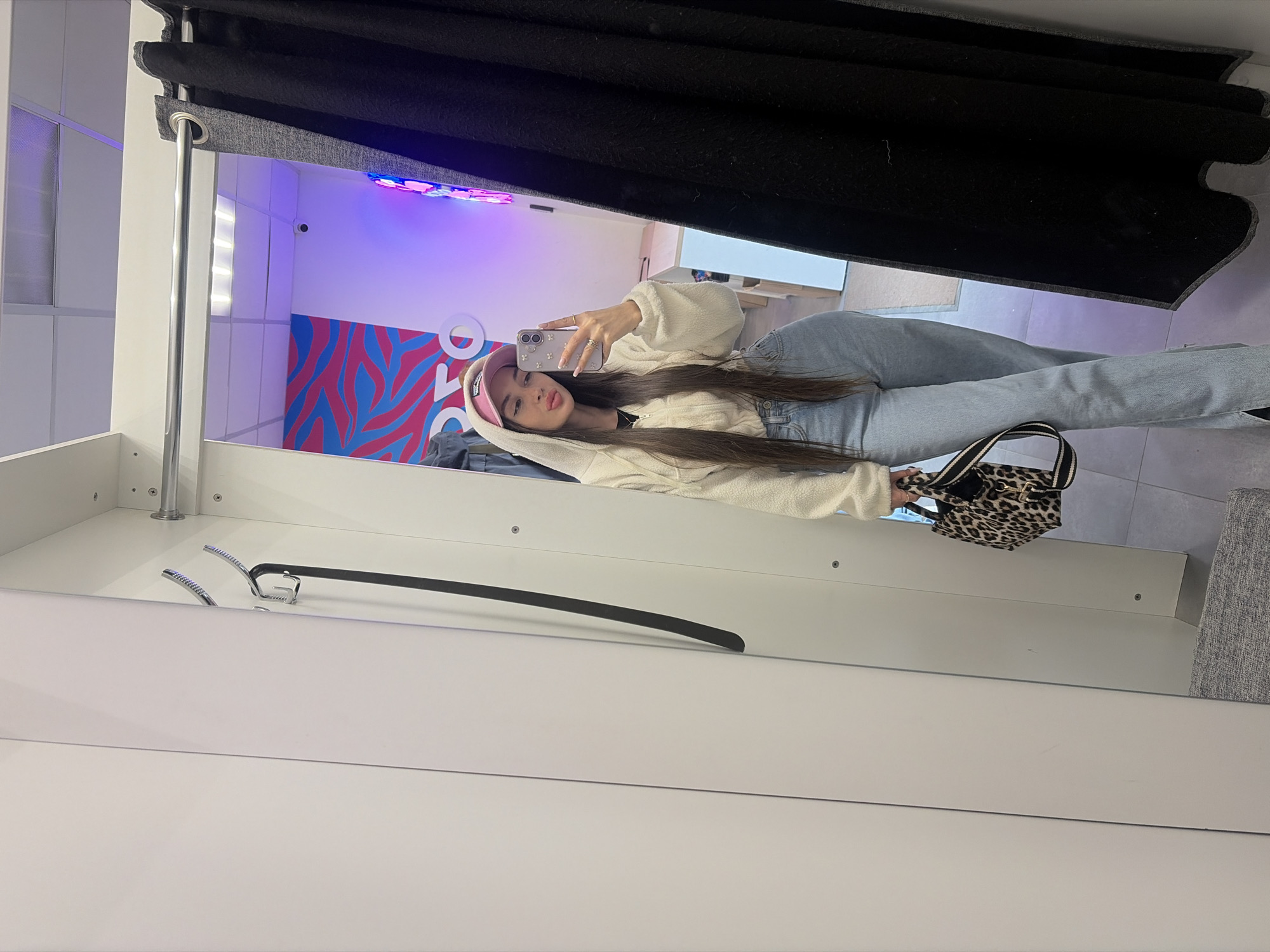 A young woman is taking a selfie in a fitting room. She is wearing a pink hat, a cream-colored hooded sweatshirt, and light blue jeans. She is holding a phone in her right hand and a small, leopard-print purse in her left hand. The fitting room has a white interior with a black curtain drawn partly across the top of the mirror. A clothes hanger is visible to the left of the woman. The background of the fitting room has a white wall with a blue and red zebra stripe design and purple LED lighting near the ceiling. This fitting room is most likely in Saint Petersburg, Russia.