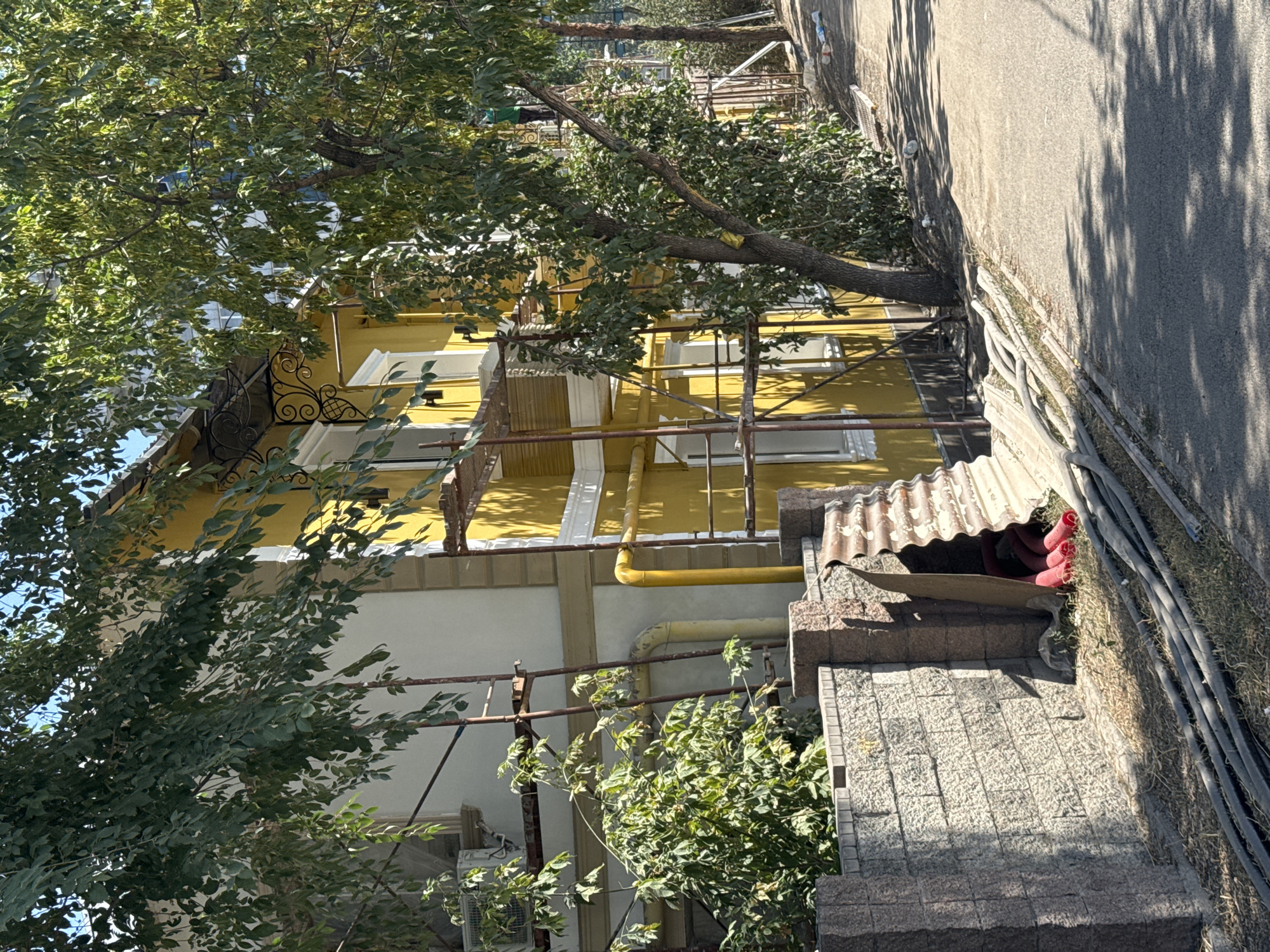 The image depicts a yellow building undergoing renovation, with scaffolding erected around its exterior. A large tree with dense foliage partially obscures the building. In the foreground, a low stone wall is visible, adjacent to a corrugated metal structure and a pile of red pipes. Several dark conduits or pipes lie on the ground in front of the wall, and the scene is bordered by an asphalt road. The overall setting suggests an urban environment. The specific location is the city of Almaty, Kazakhstan.