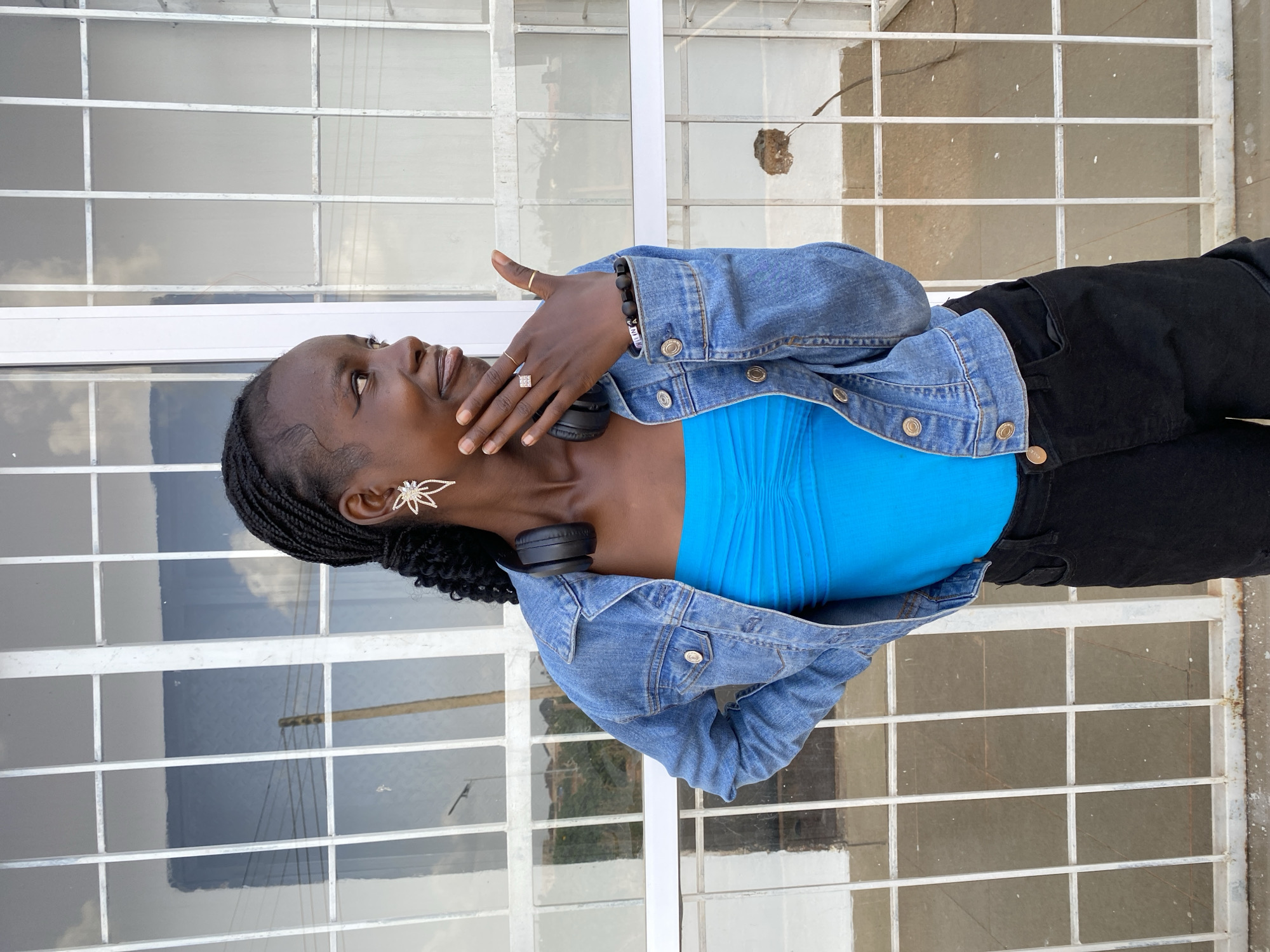 A young Black woman stands in front of a white-barred window, looking to her right. She has braided hair and is wearing a bright blue strapless top, a denim jacket, and black pants. Her left hand is raised to her face, touching her cheek, and she is wearing rings on multiple fingers. On her right ear, she has a star-shaped earring with a pearl at the center. A pair of black headphones rests around her neck. She is also wearing beaded bracelets on her left wrist. The window behind her has a reflection of the sky and some faint outlines of buildings or structures. The ground appears to be paved. The location is specified as Bar Jirgi Summa, Nigeria, which implies an outdoor setting in that region.