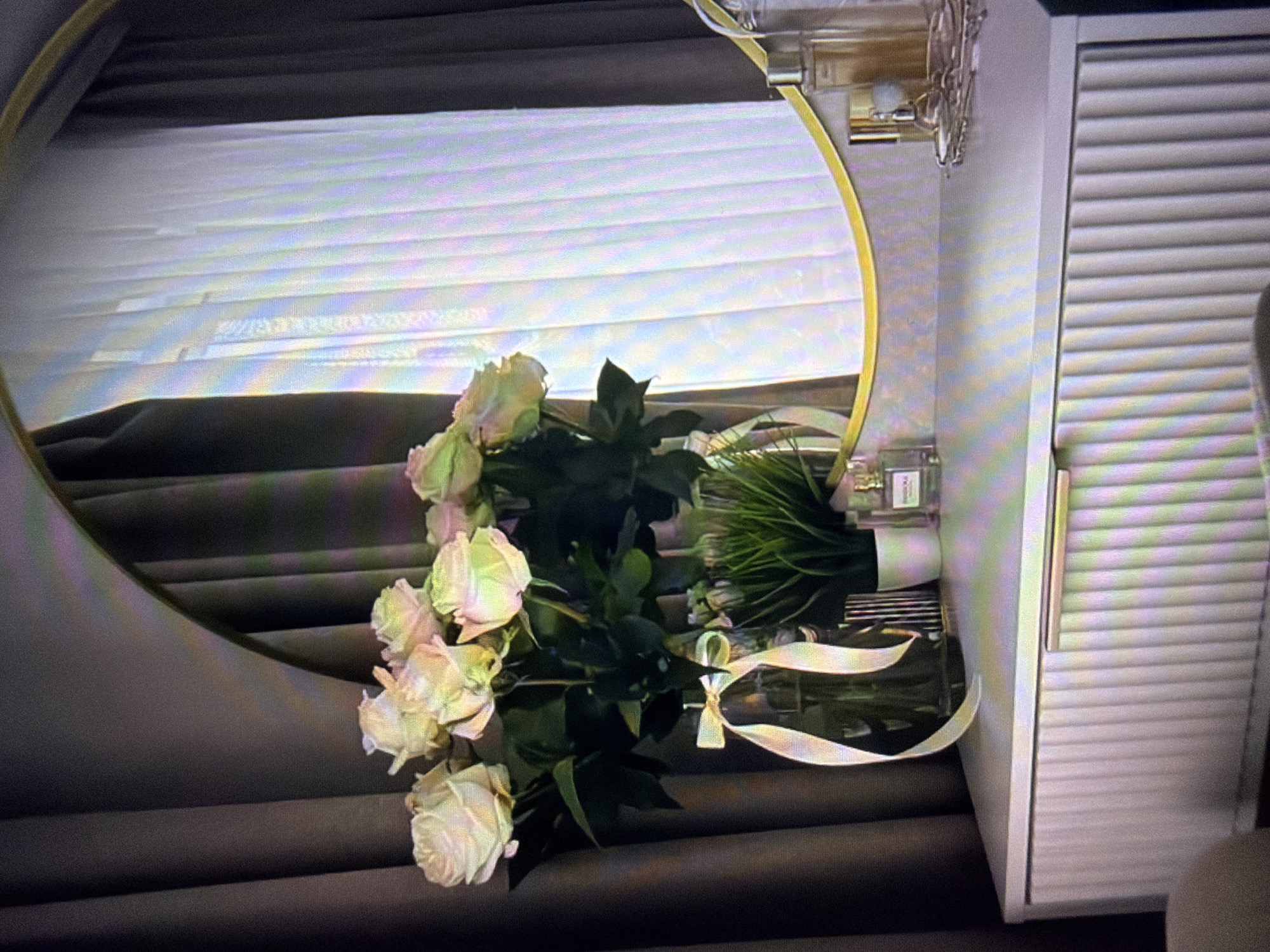 This is a close-up, horizontal, medium-shot photograph of a bouquet of pale pink roses arranged in a glass vase filled with water and greenery. The bouquet is placed in front of a large, round mirror with a thin gold frame. The mirror reflects the scene, including a set of blinds in the background which are partially obscured by the bouquet and its reflection. To the right of the mirror, a white cabinet with horizontal slatted doors is visible. Two perfume bottles are also visible on a surface behind the bouquet and in the mirror's reflection. One bottle is clear with a silver cap, and the other appears to be a dark green glass bottle. A pale ribbon is tied around the neck of the dark green bottle, and a similar ribbon is tied into a bow on the bouquet itself, draped over the vase. The overall lighting suggests an indoor setting, likely during daylight hours due to the reflection of the blinds. The scene evokes a sense of quiet elegance and domesticity. There is no visible text in the image.