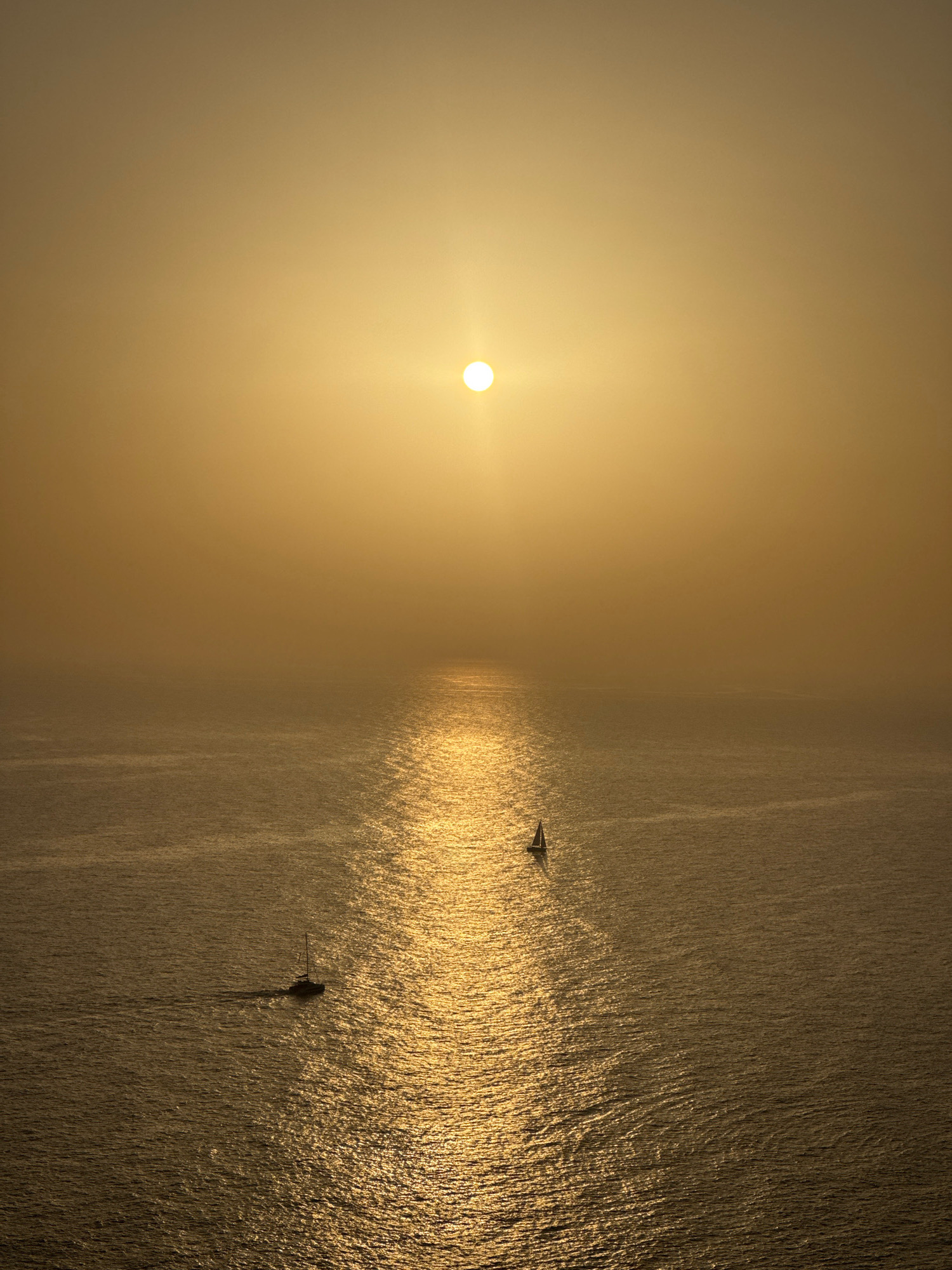 The image depicts a serene ocean scene at sunset. The sun, a bright orb, is positioned high in the hazy, golden sky, casting a shimmering path of light across the water. Two sailboats are visible on the water, one further away and closer to the center of the sun's reflection, and the other nearer to the bottom left of the frame, moving away from the viewer. The water itself is calm with gentle ripples that catch the sunlight. The overall atmosphere is tranquil and bathed in warm, golden hues. This scene is located in the city of Zagora, Montenegro.