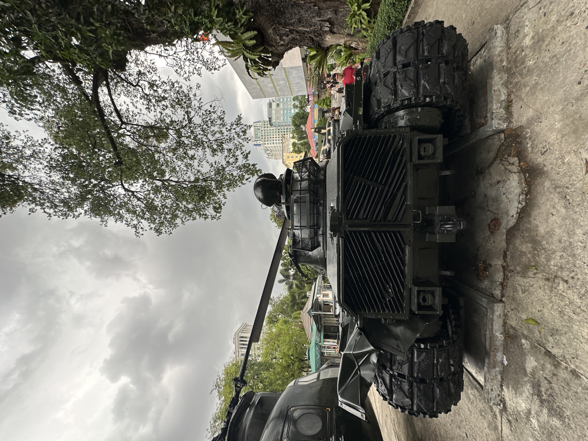 The image shows a Soviet-era tank on display in the city of Thủ Đức, Vietnam. The tank is positioned on a concrete pad, with a tree and some shrubbery in the background. People are visible near the back of the tank. Buildings and cloudy skies are visible in the background. The tank's tracks, radiator, and part of the turret are visible.