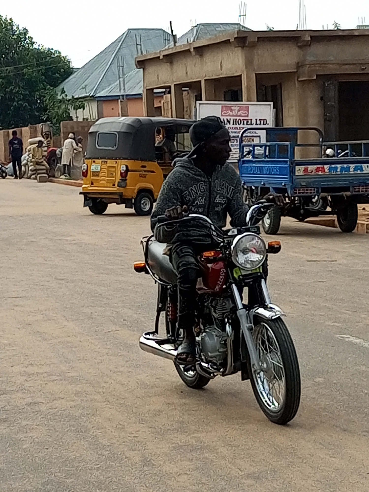 A man wearing a dark baseball cap and a grey hooded sweatshirt is riding a motorcycle on a paved street in Tempure, Nigeria. The motorcycle is red and silver with spoked wheels. The man is looking forward and appears to be the sole occupant of the motorcycle. In the background, there is a yellow auto-rickshaw, a blue truck with "ROYAL MOTORS" and "LALAMMA" written on it, and several buildings. There are also piles of what appear to be sacks of goods on the side of the street. The sky is overcast.