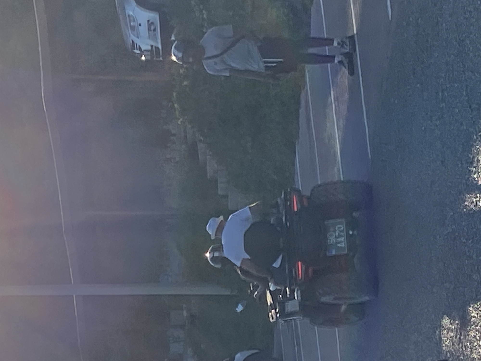 In Prijevor, Montenegro, two individuals are present in a parking lot. The foreground features an all-terrain vehicle (ATV) with two occupants, both wearing helmets. The driver of the ATV is wearing a white t-shirt and a bucket hat. The passenger is mostly obscured, but appears to be wearing a helmet. The ATV has a license plate visible that reads "BD AA70". To the right of the ATV, a second individual, also wearing a helmet and a light grey t-shirt with a dark crossbody bag and dark shorts, stands facing away from the camera. In the background, a white car is parked under a covered structure. The environment appears to be outdoors with vegetation visible behind the parked car. The lighting suggests it is either early morning or late afternoon.