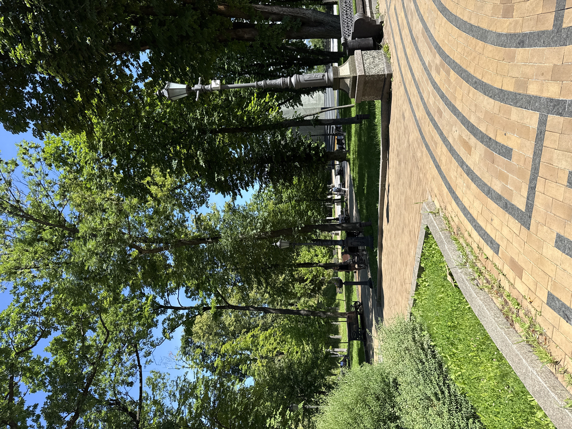 This is a photograph of a park pathway in Kyiv, Ukraine, on a sunny day. The pathway is paved with tan and dark grey interlocking bricks arranged in a curved pattern. To the left of the pathway is a raised garden bed filled with green grass and some weeds, bordered by a stone wall. Adjacent to this is a dense green bush. Further into the park, more paved paths and green grass areas are visible, dotted with benches and lamp posts. Several mature trees with lush green foliage line the park, casting dappled shadows on the ground. In the background, a few people can be seen in the distance, walking or sitting on benches. The sky is a clear, bright blue.