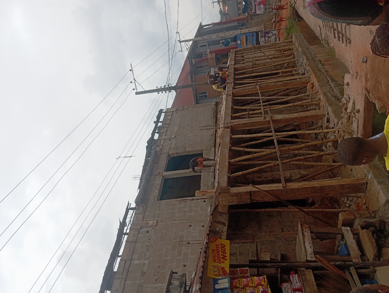 The image captures an active construction site for a multi-story building in Nkpor, Nigeria, under an overcast sky during daytime. The building, constructed with concrete blocks, shows rebar protruding from its upper levels, indicating ongoing work. Extensive scaffolding, a mix of wooden planks, sawn timber, and bamboo poles, supports an unfinished upper floor or roof slab.
Several people are visible, indicating activity. Two workers are on the upper levels of the building, with one person in an orange shirt clearly seen on a concrete slab. Other individuals are further up on the scaffolding structure. In the immediate foreground on the right, partial views of onlookers or passersby are visible, including a person in a yellow shirt and another's head.
To the right of the construction, the ground appears to be a dirt path or undeveloped area strewn with rubble and building materials. In the background, other completed buildings of similar construction line what seems to be a street or residential area. Overhead power lines are strung across utility poles against the cloudy sky.
In the bottom left foreground, a small, makeshift stall or shop is set up beside the construction site, displaying various packaged goods. A yellow sign attached to this stall prominently displays the text "MALTA GUINNESS N600," likely indicating the price of Malta Guinness in Nigerian Naira. The scene reflects typical urban development and daily life in the region.
