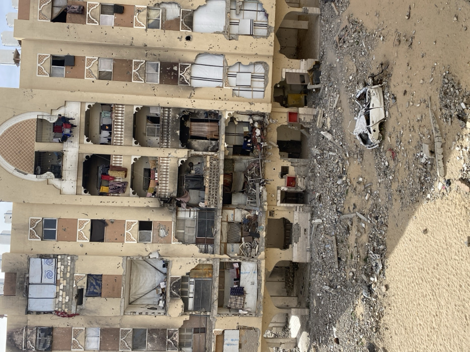 The image depicts a multi-story residential building in Khan Yunis, Palestinian Territory, exhibiting extensive damage. The light-brown facade of the building is perforated by numerous circular projectile impact holes and shrapnel marks. Large sections of external walls and window frames are missing across multiple floors, exposing the interiors of at least seven apartment units. Balconies display structural damage; some contain visible items, including laundry hanging on lines, while others show signs of charring and fire damage. Makeshift coverings, such as white tarpaulins and corrugated metal sheets, are used to cover some exposed openings. The ground directly in front of the building is a debris field consisting of pulverized concrete, twisted metal, and other rubble, interspersed with sandy patches. A severely damaged, white four-door vehicle, largely collapsed and missing its roof and significant body panels, is situated amidst the debris in the mid-right portion of the foreground. No individuals are present in the scene.