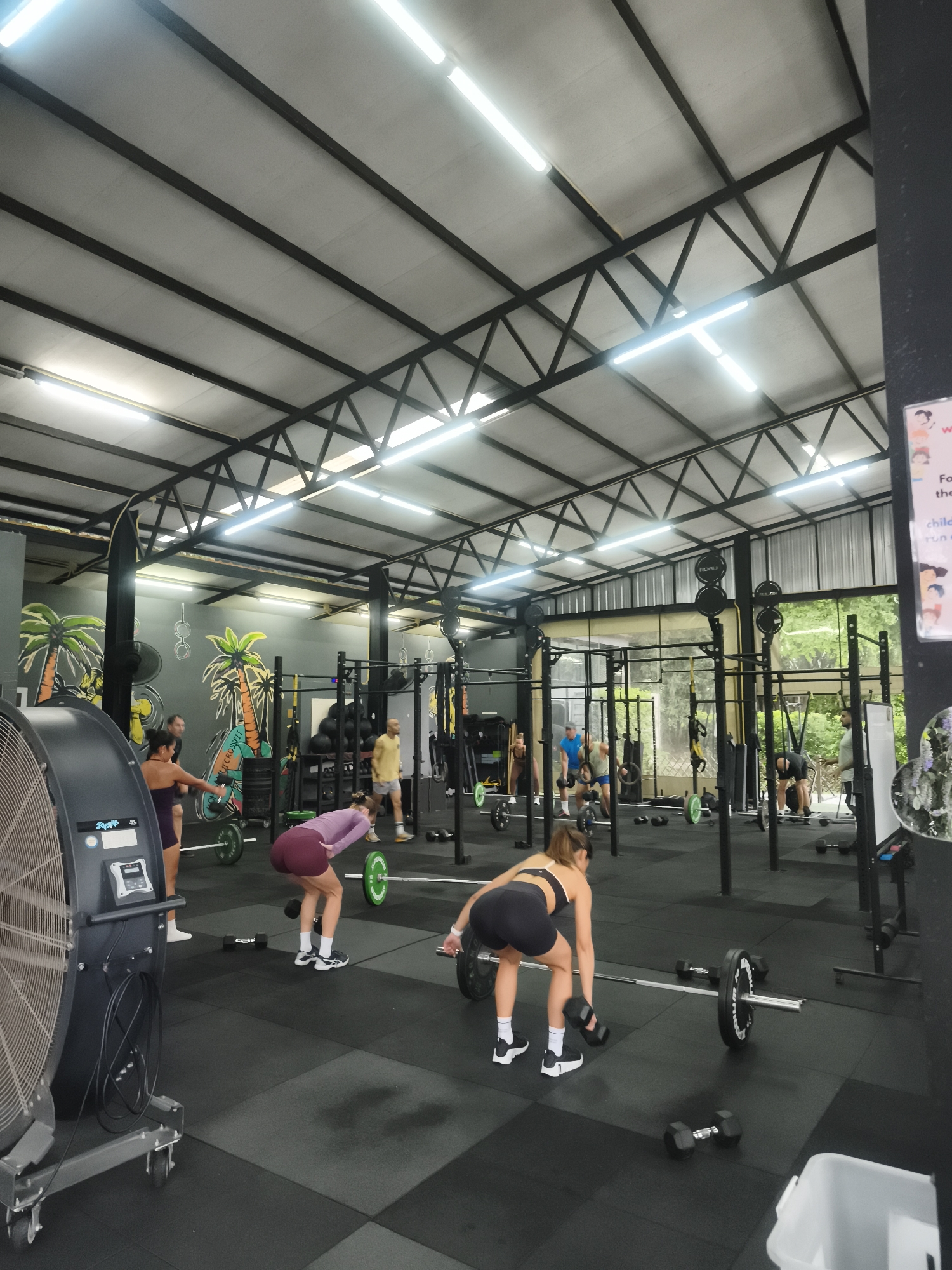 The image shows a CrossFit gym in Baan Bo Phut, Thailand. The gym has a high ceiling with exposed metal beams and bright lighting. Several people are engaged in various exercises. Some are lifting weights with barbells and weight plates, while others are using equipment like pull-up bars and gymnastic rings. A large fan stands on the left side of the frame. The floor is covered in black rubber mats. The walls are decorated with tropical-themed murals featuring palm trees. The overall atmosphere is active and focused on physical fitness.