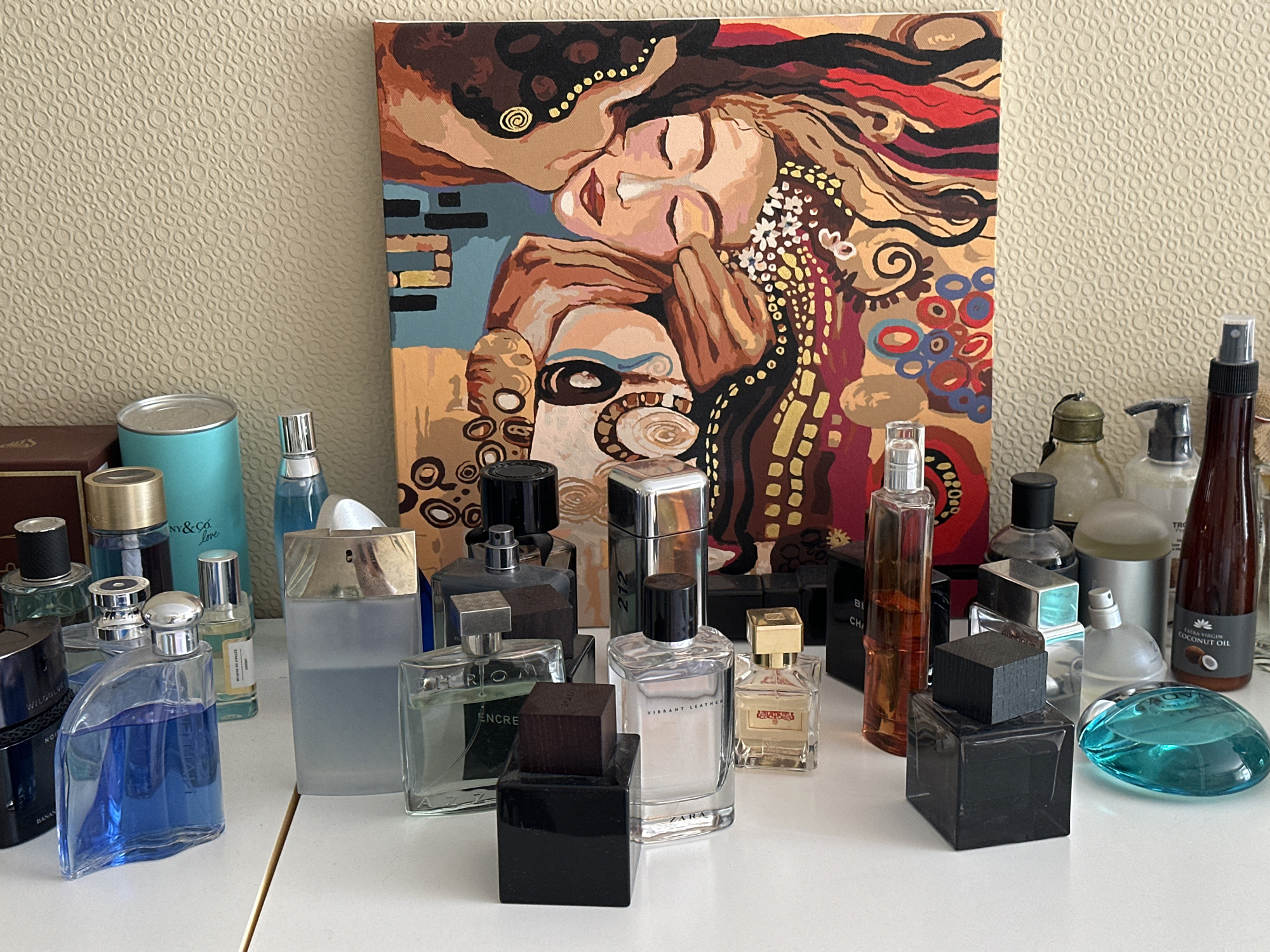 The image displays a collection of perfumes and a framed artwork on a white surface, possibly a dresser or shelf. The artwork, a painting with abstract figures and bold colors, is positioned behind the perfume bottles. The perfumes vary in size, shape, and color, with some clear bottles showcasing blue or clear liquids, while others contain amber or dark liquids. One prominent bottle is a large, oval-shaped teal glass perfume bottle. The background features a textured, light-colored wall. The scene is well-lit, likely from overhead lighting, casting soft shadows on the objects. There are no people visible in the image. The specific location in the city of Nevinnomyssk, Russia, is not discernible from the provided image.