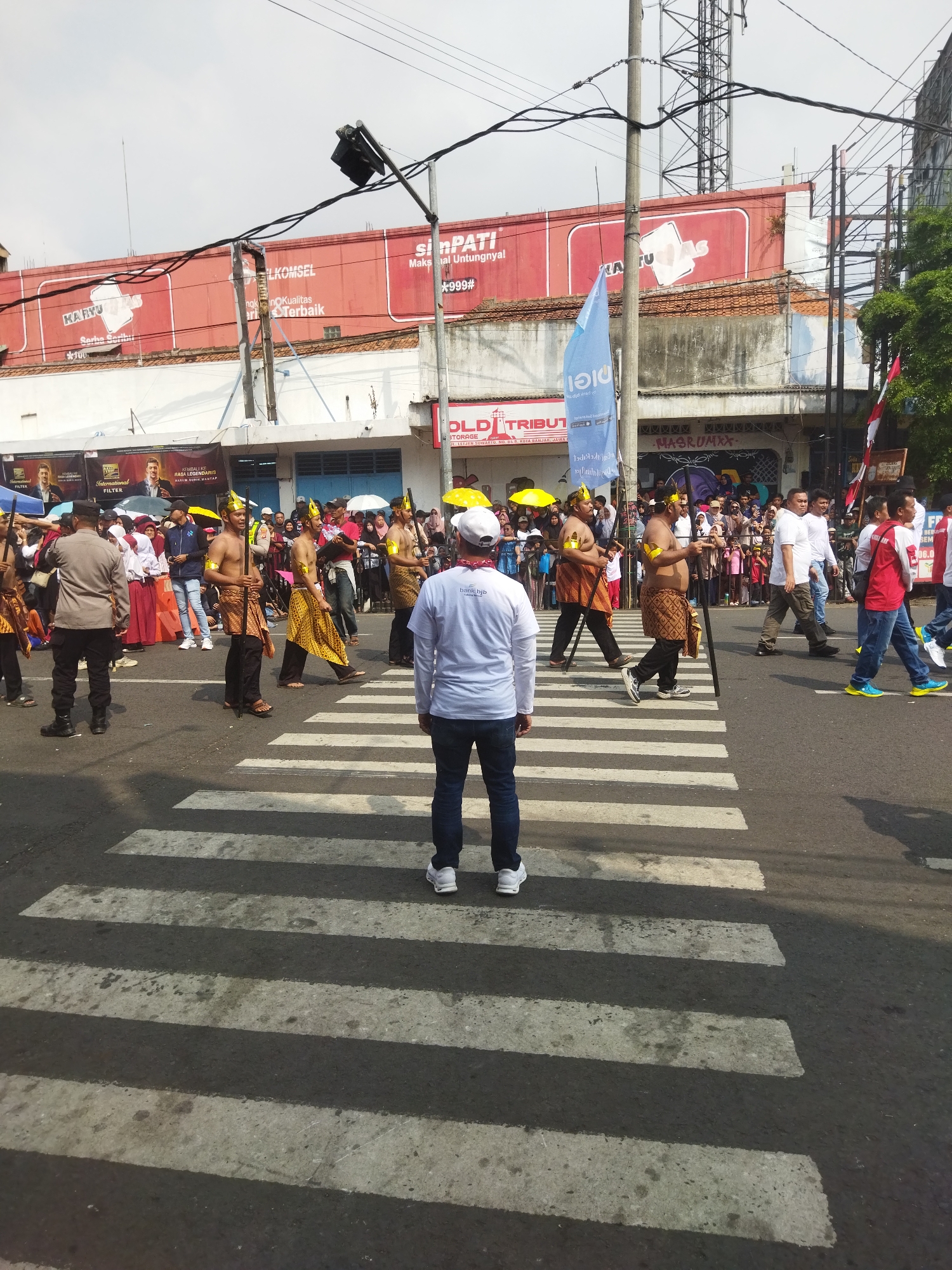 On a bright, overcast day, a parade is taking place on a city street in Banjar, Indonesia. A group of men in traditional attire, consisting of loincloths and headwear, march across a pedestrian crossing. They are armed with staffs. In the foreground, a man in a white t-shirt with the text "bank bjb" and dark blue jeans stands facing away from the camera, also on the pedestrian crossing. Behind the parading men, a crowd of spectators lines the street, some holding umbrellas. Buildings with red awnings and various signs, including "TELKOMSEL," are visible in the background, along with a large communication tower. The scene suggests a cultural event or celebration occurring in the urban environment of Banjar.